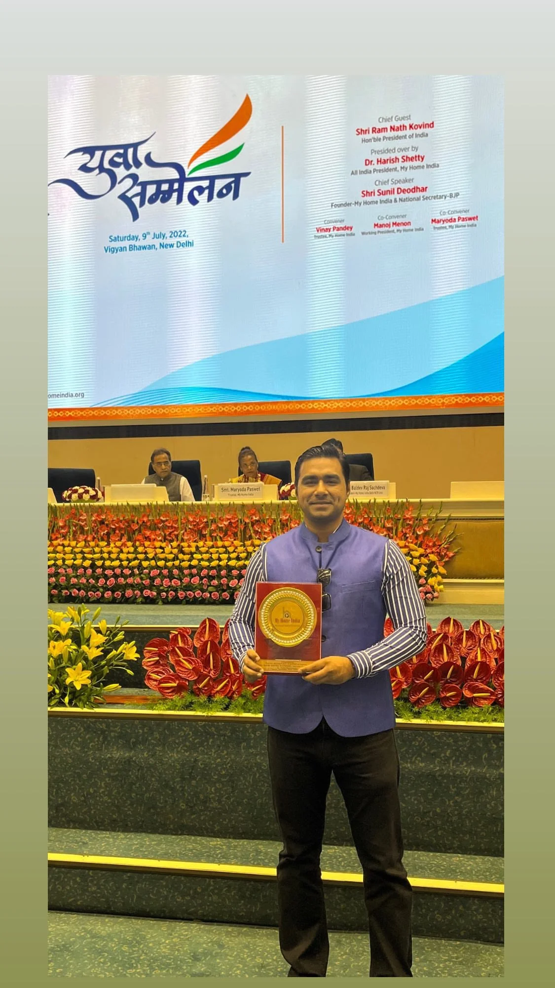 A man standing on stage holding a plaque at an event in India, with a large screen behind him displaying event details and three individuals seated at a table.