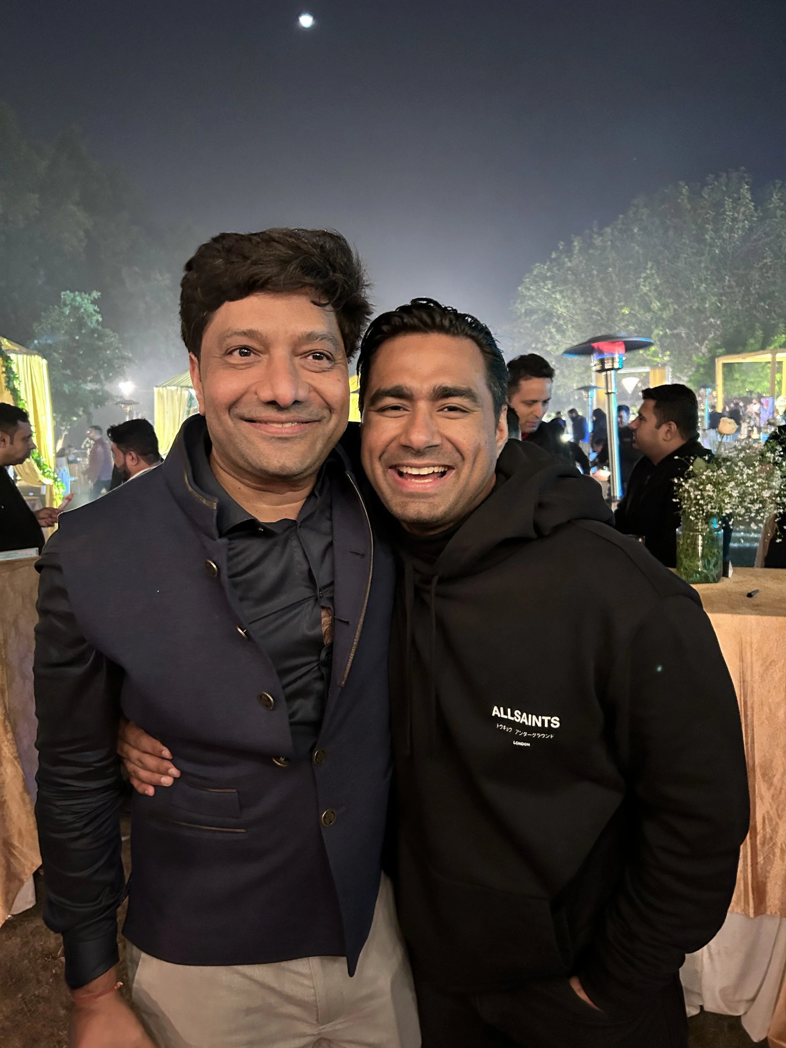 Two men smiling and posing together at an outdoor event during nighttime. The background features trees, decorative lighting, and people socializing.