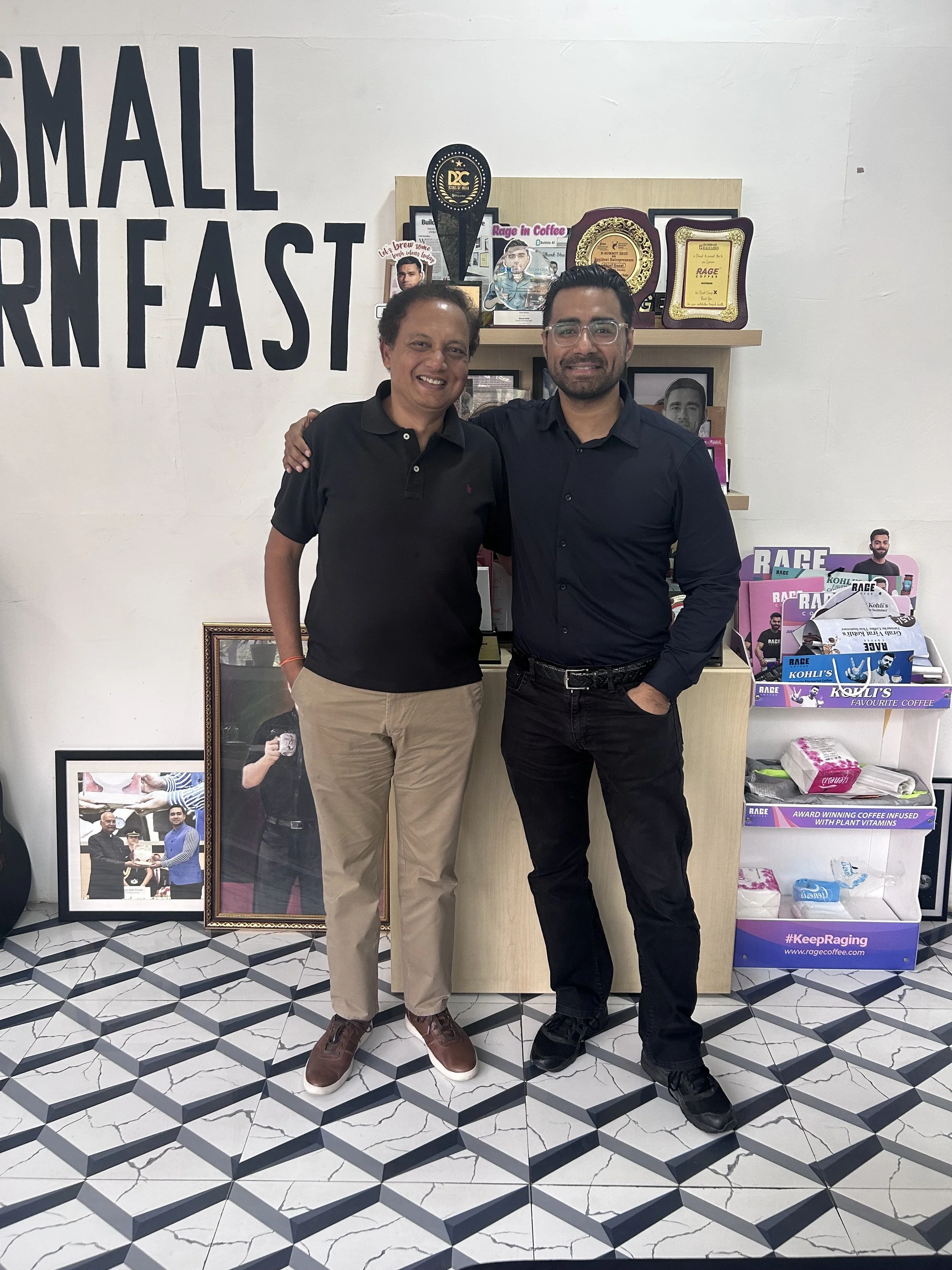Two men standing side by side indoors, smiling at the camera. One is wearing a black polo shirt and khaki pants, the other is wearing a dark blue dress shirt and black pants. They are in front of a shelf with awards, plaques, and coffee products, wit
