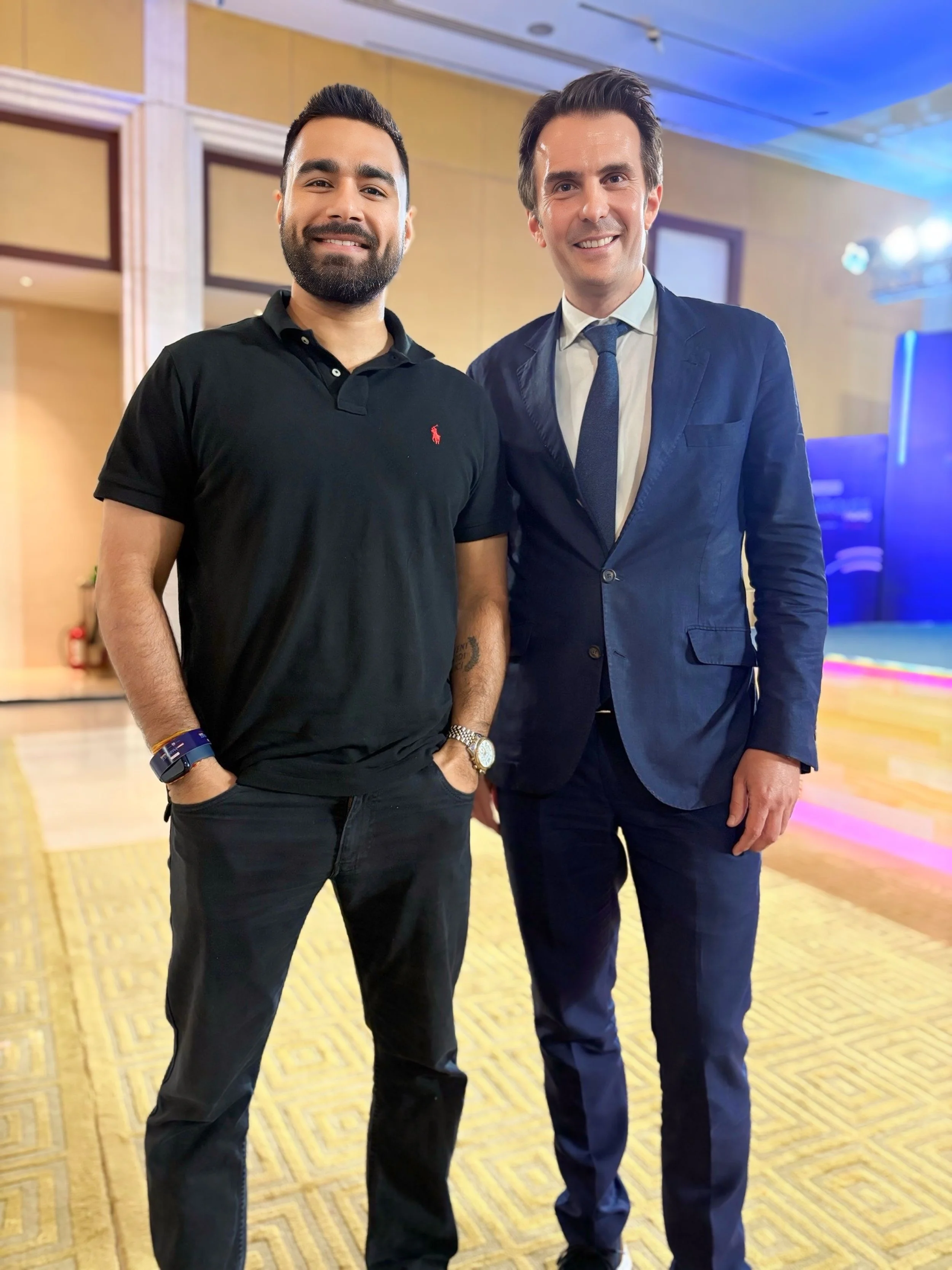 Two men standing together, smiling for the camera indoors. One man is dressed casually in a black polo shirt and black pants, the other is in a dark blue suit with a white shirt and tie. The background shows a decorated stage area with lighting.