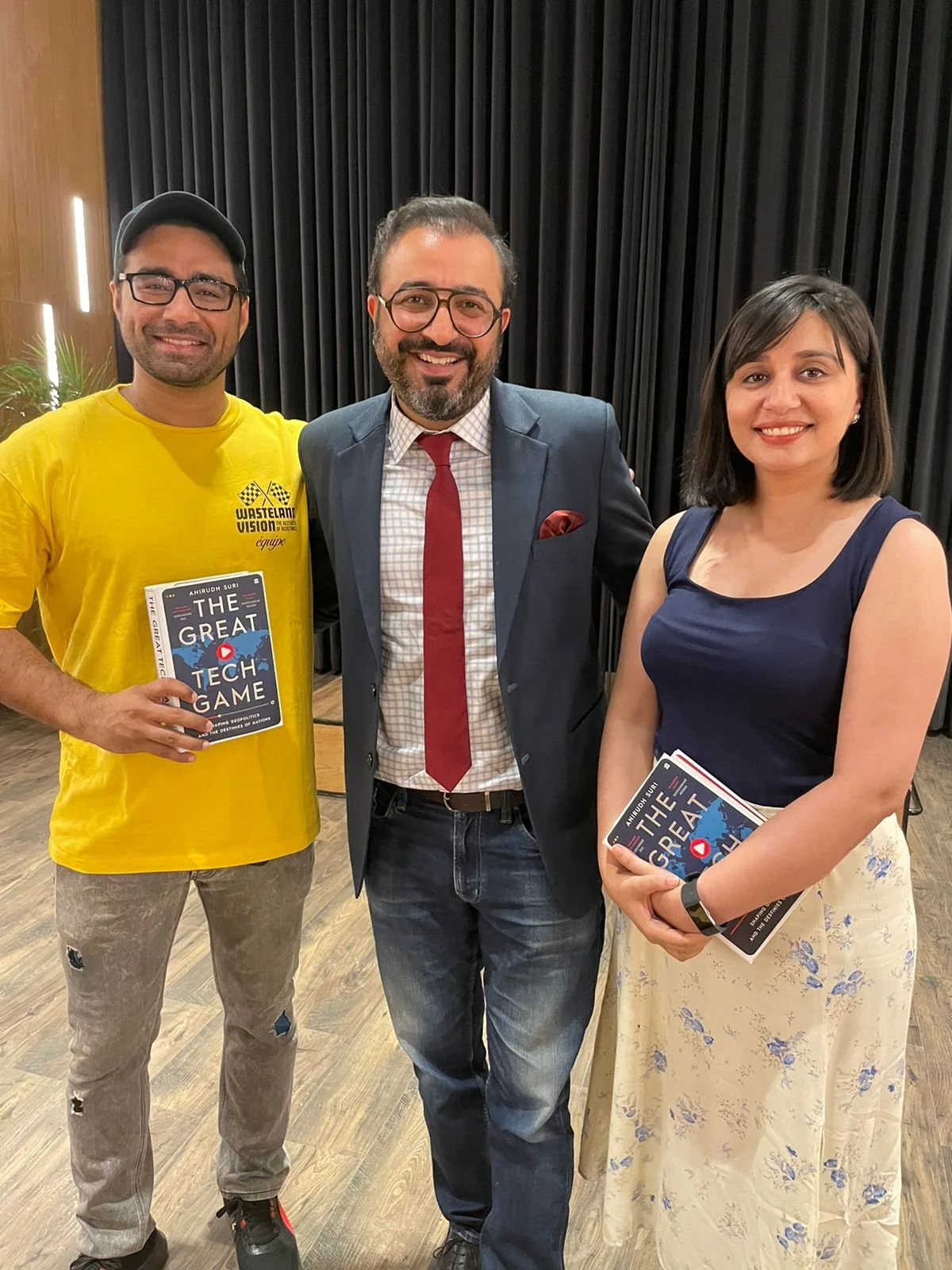 Three people standing together indoors, smiling. The man on the left is wearing a yellow t-shirt, glasses, and a cap, holding a book titled 'The Great Tech Game.' The man in the middle is dressed in a suit with a red tie, glasses, and has a beard. Th