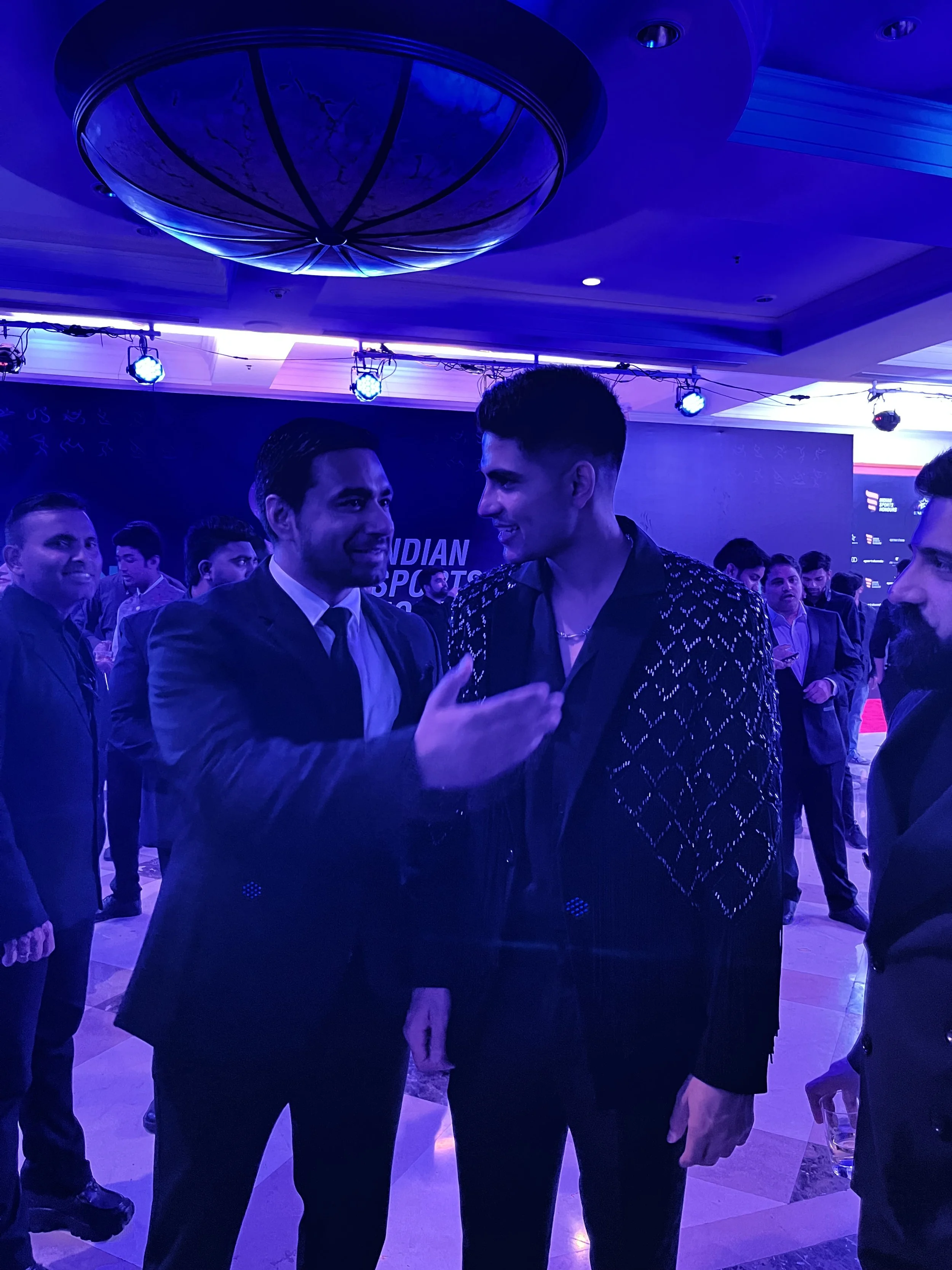 Two men in conversation at an event with a crowd in the background, under blue lighting and ceiling decor.