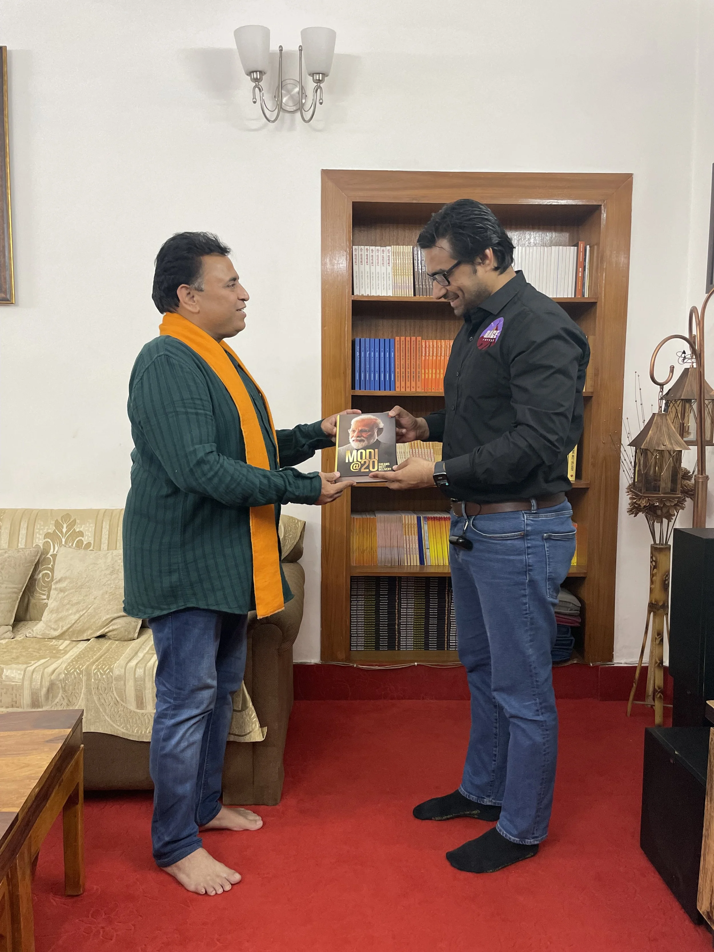 Two men exchanging a book in a living room with a couch, a small wooden table, and a bookshelf in the background. One man is barefoot and wearing a green and orange jacket, the other is wearing glasses and blue jeans. They are smiling and standing on
