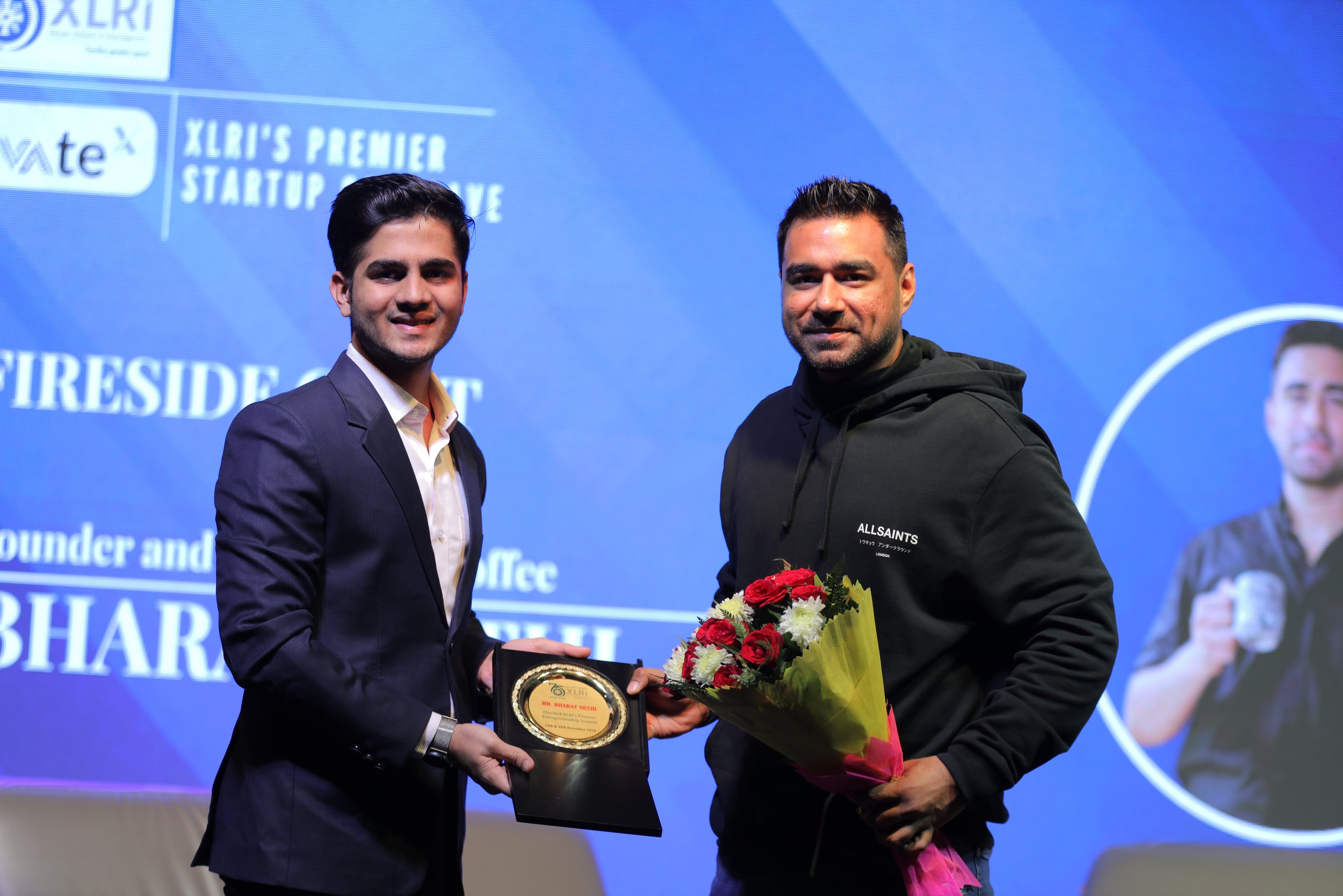 Two men standing on stage, one in a suit and the other in a black hoodie, holding a plaque and a bouquet of flowers, with a blue event backdrop in the background.