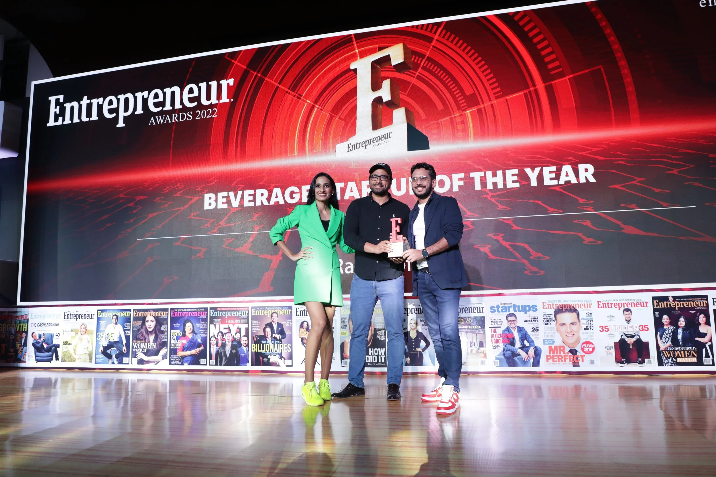 Three people standing on a stage, holding an award at the Entrepreneur Awards 2022. Behind them is a large digital screen displaying 'Entrepreneur AWARDS 2022' and 'BEVERAGE STARTUP OF THE YEAR'.