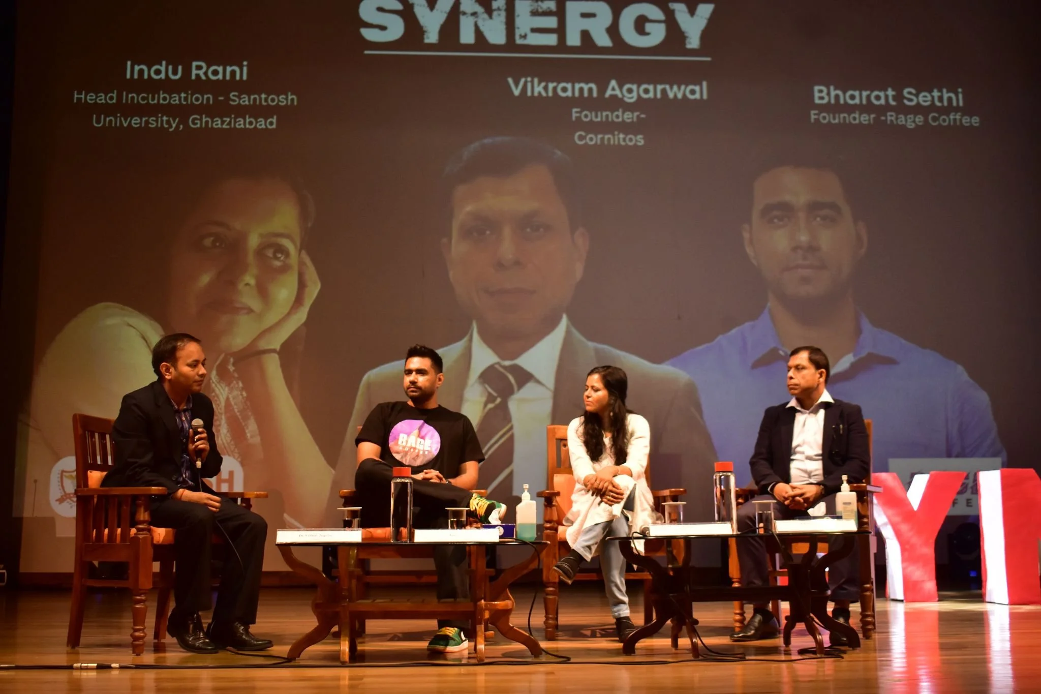Panel discussion with four individuals seated on stage, two men and two women, talking into microphones. Behind them, a large screen displays images of three other people with names and titles. The stage has a wooden floor with tables, water bottles,