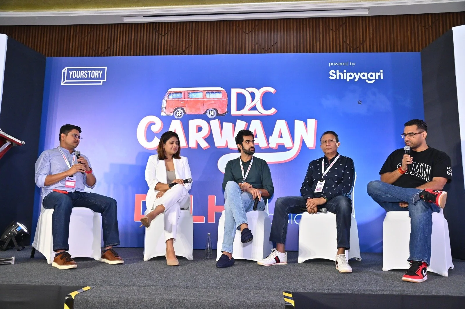 Five people seated on a stage at a panel discussion, with a blue backdrop displaying the words "D2C CarWaan" and logos for "YourStory" and "Shipyaari." The panelists are holding microphones, engaging in conversation.