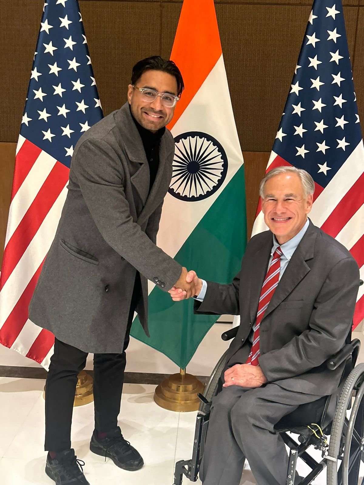 Two men shaking hands in front of American flags and the Indian flag, with one man in a wheelchair.