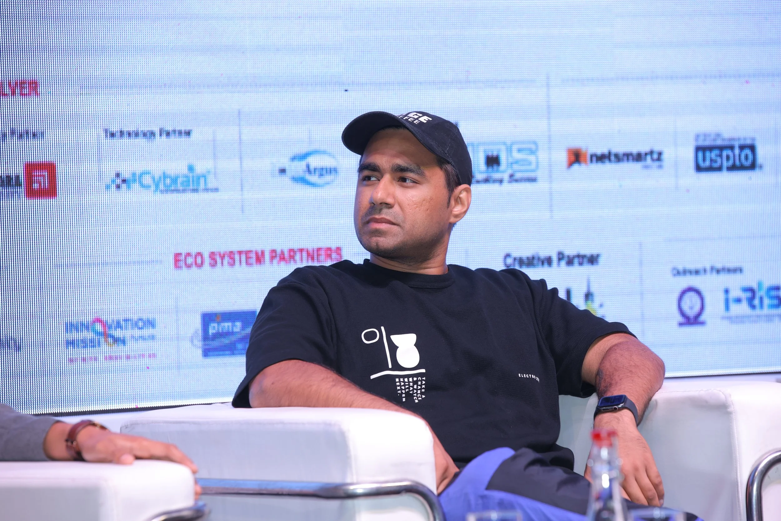 A man in a black t-shirt and black cap sitting on a white couch at a conference with a large screen behind him displaying various sponsor logos.