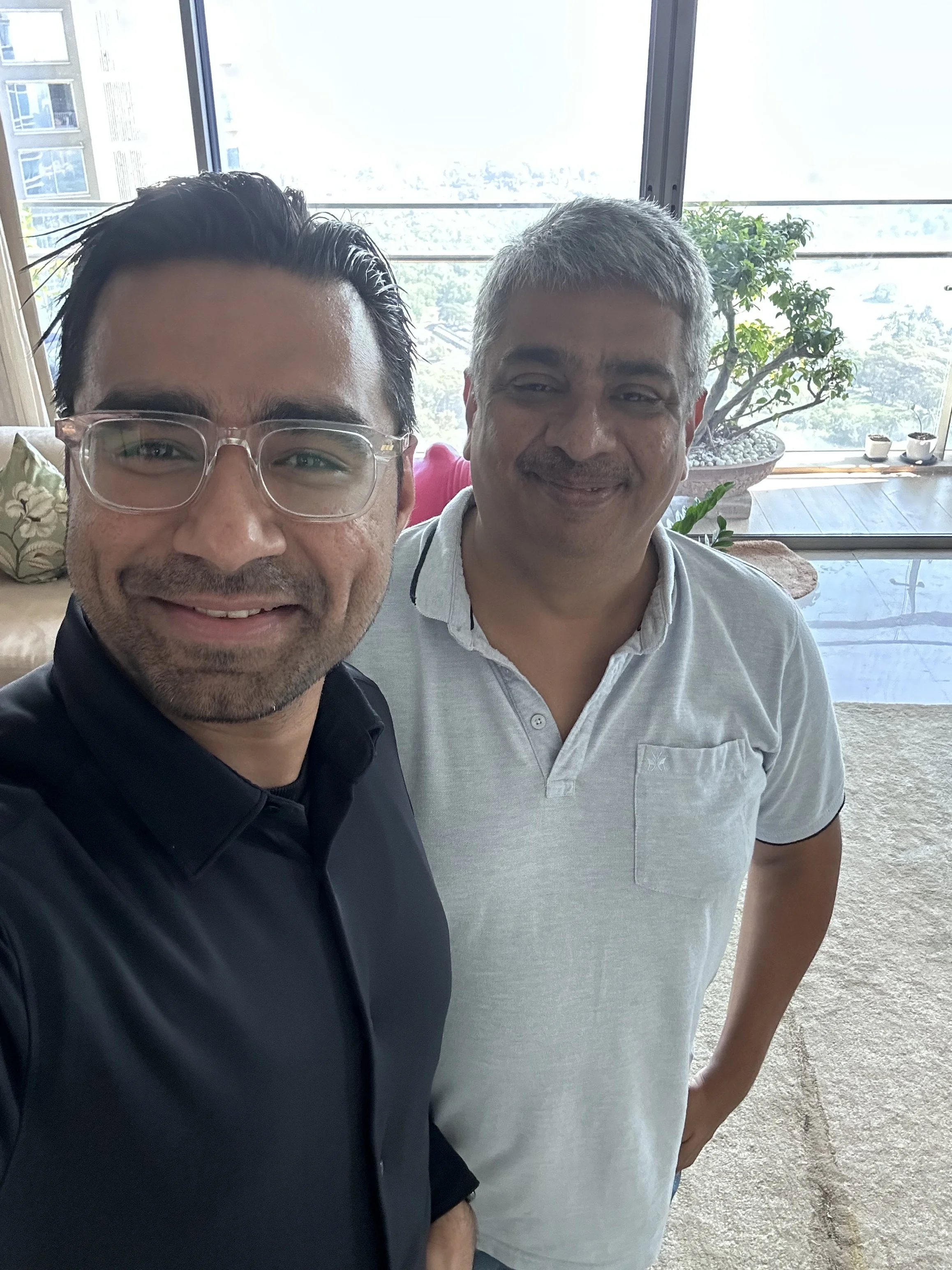 Two men taking a selfie together in a bright room with large windows showing trees and buildings outside.