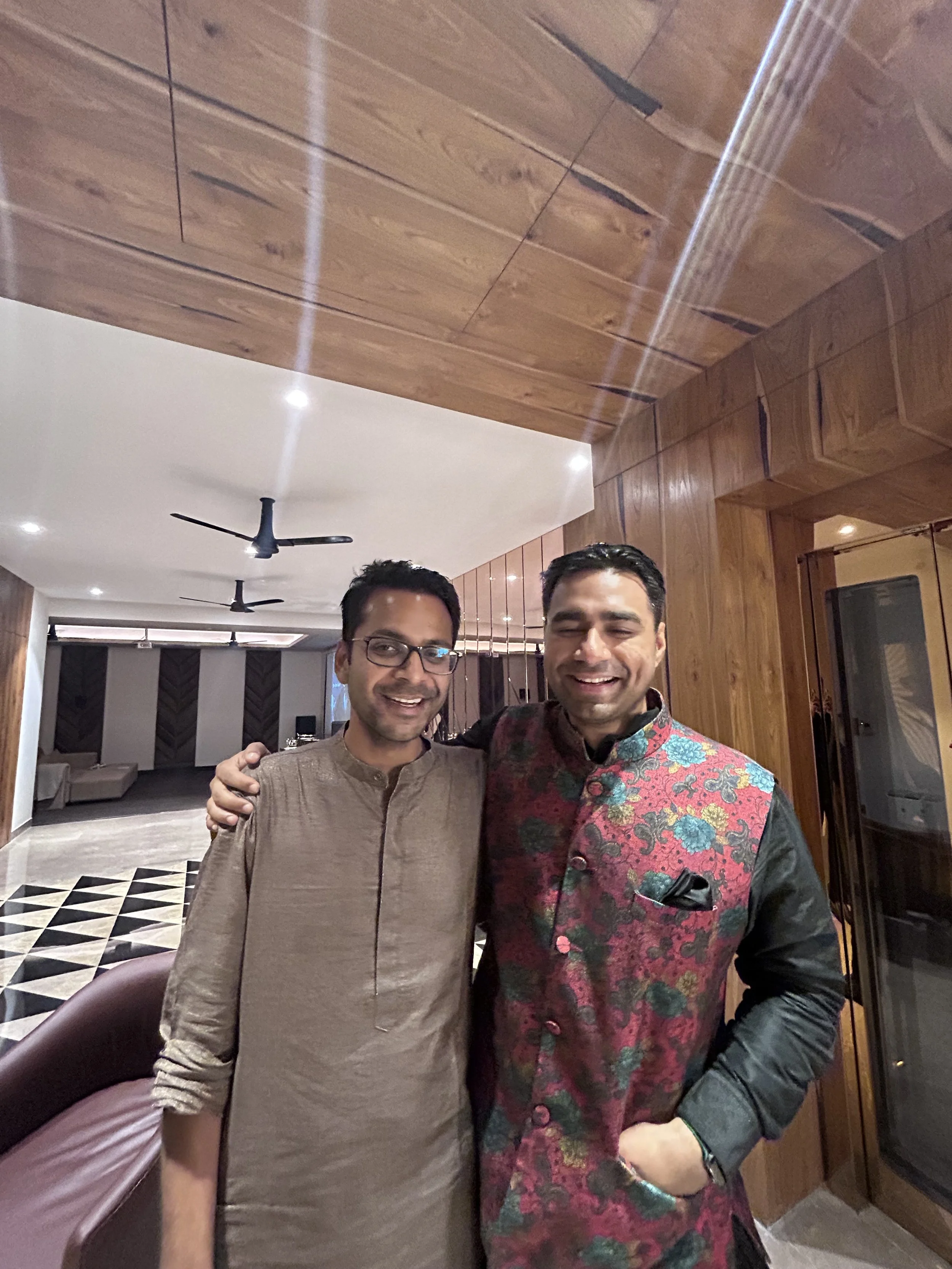 Two men smiling and posing together indoors, one wearing glasses and a brown kurta, the other in a floral vest over a black long-sleeve shirt, with wooden wall and ceiling, ceiling fans, and modern decor in the background.