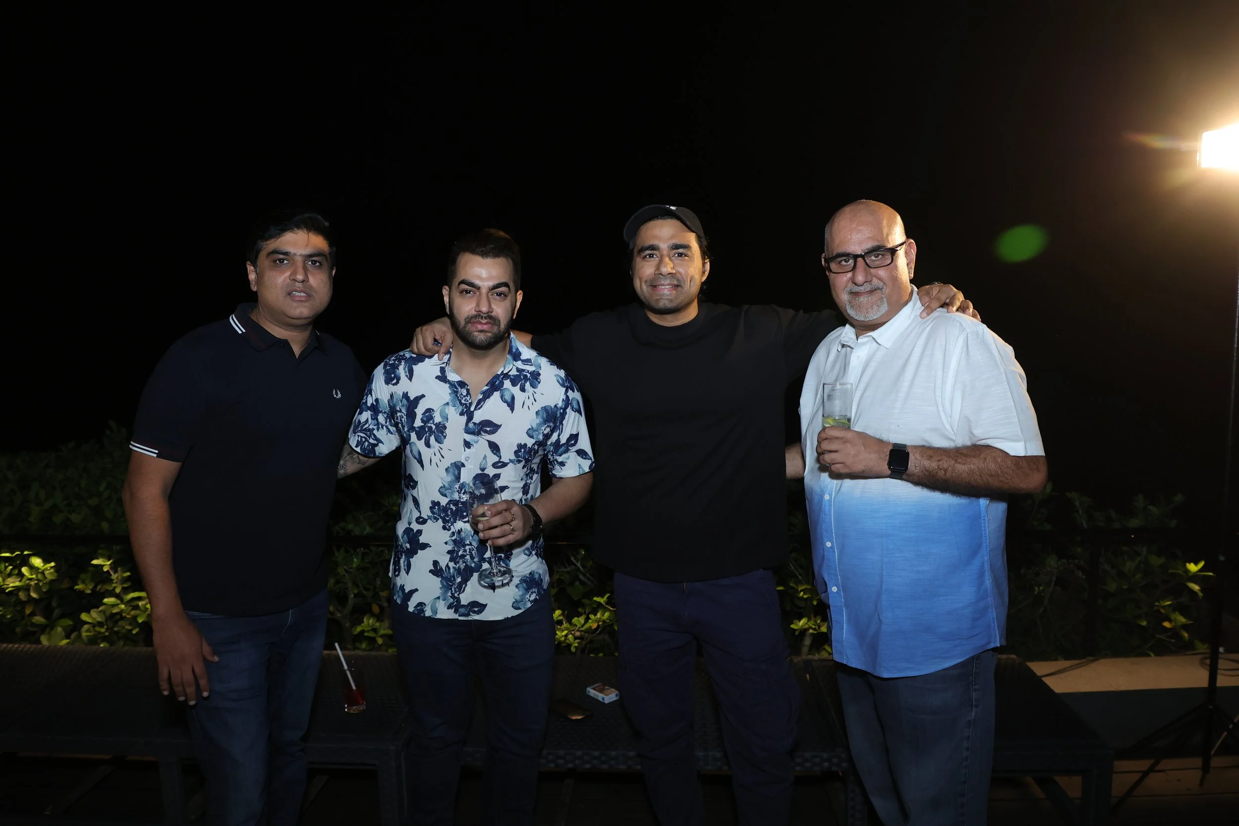 Four men standing together at night, smiling, with one holding a drink, and a dark sky in the background.