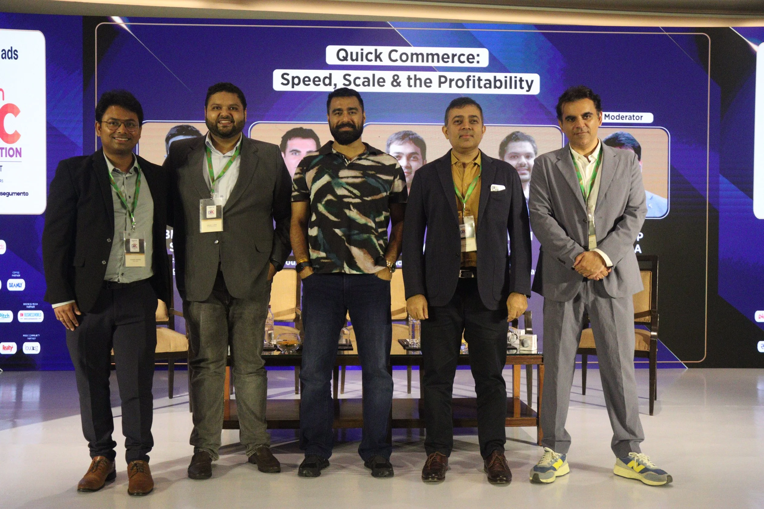 Five men standing in front of a presentation screen at a conference, smiling for the photo. The screen displays the title 'Quick Commerce: Speed, Scale & the Profitability' and a panel layout with a moderator.