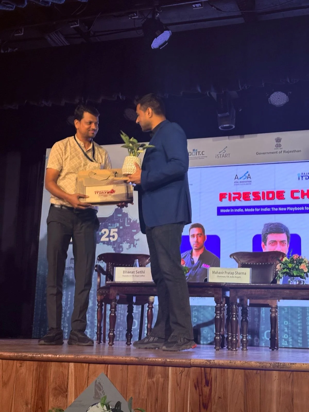 Two men on stage at an event, one presenting a potted plant and a box to the other. Behind them is a large screen with text and images of two men, and there are chairs with nameplates in front of the screen.