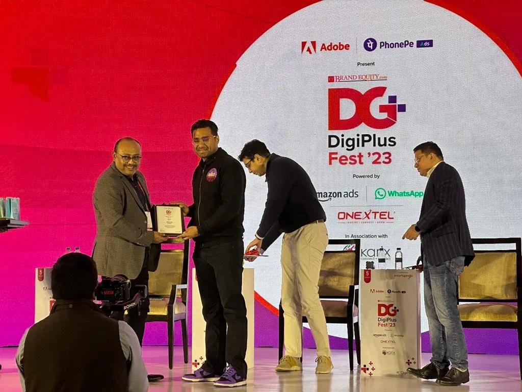 Two men on stage during an award ceremony at DigiPlus Fest 23, with one presenting a framed certificate or award to the other. Two other men are on the stage, one looking at a mobile device and the other standing with his hands in his pockets. A came
