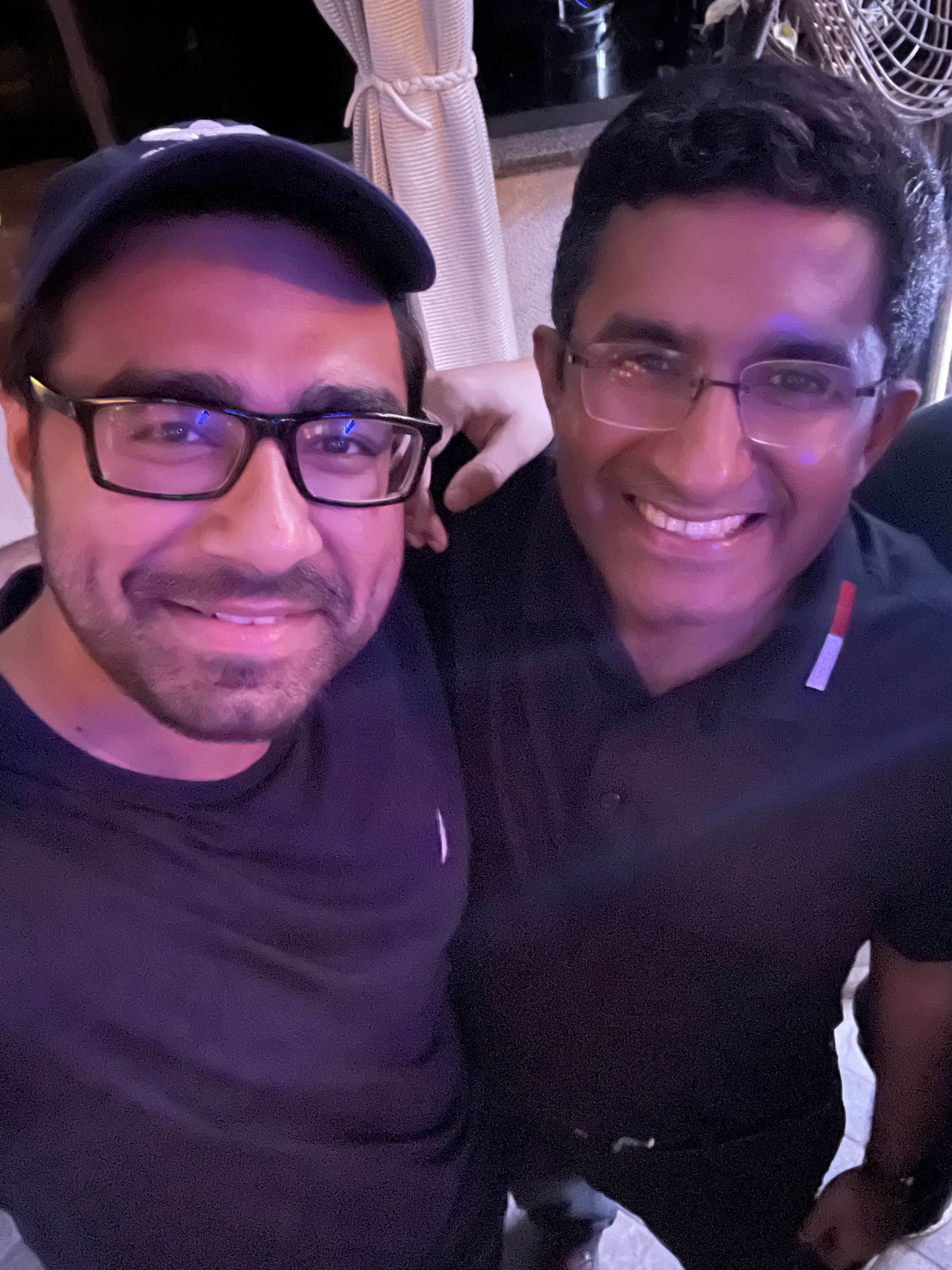Two men smiling for a selfie indoors, with curtains and a fan visible in the background.