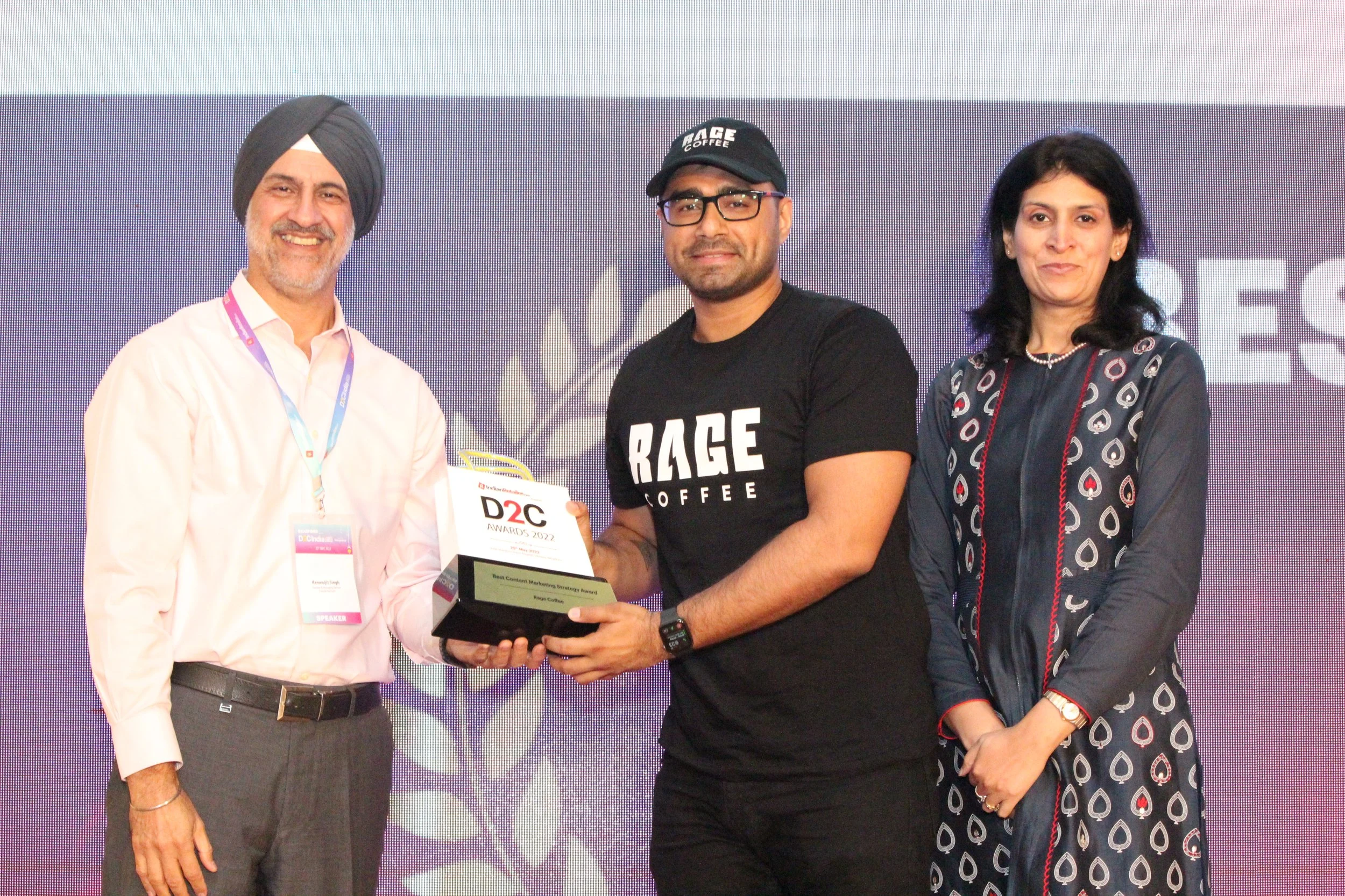 Three people on stage at an awards ceremony, with one man in a white shirt and turban holding a trophy, another man in a black technology T-shirt and glasses receiving the trophy, and a woman in a patterned dress standing next to them, all smiling. T