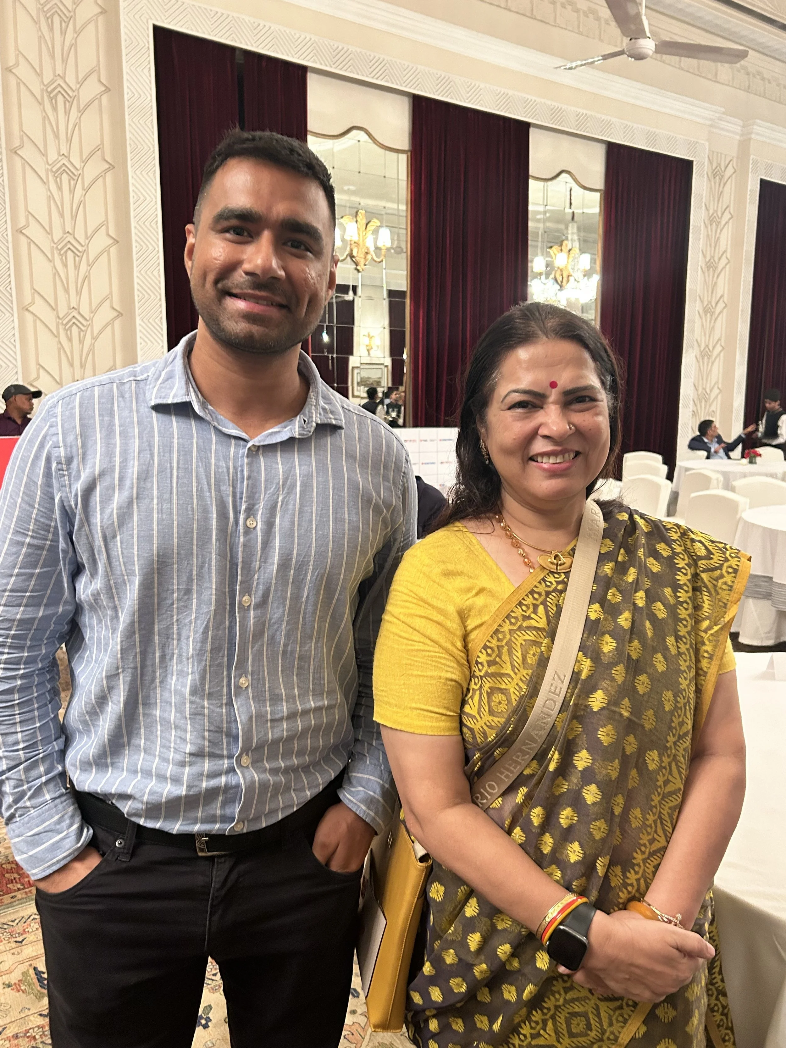 A man and a woman standing side by side in a banquet hall, smiling at the camera. The man is wearing a light blue striped shirt and dark pants, with hands in pockets. The woman is dressed in a traditional yellow and brown sari with gold accents, wear