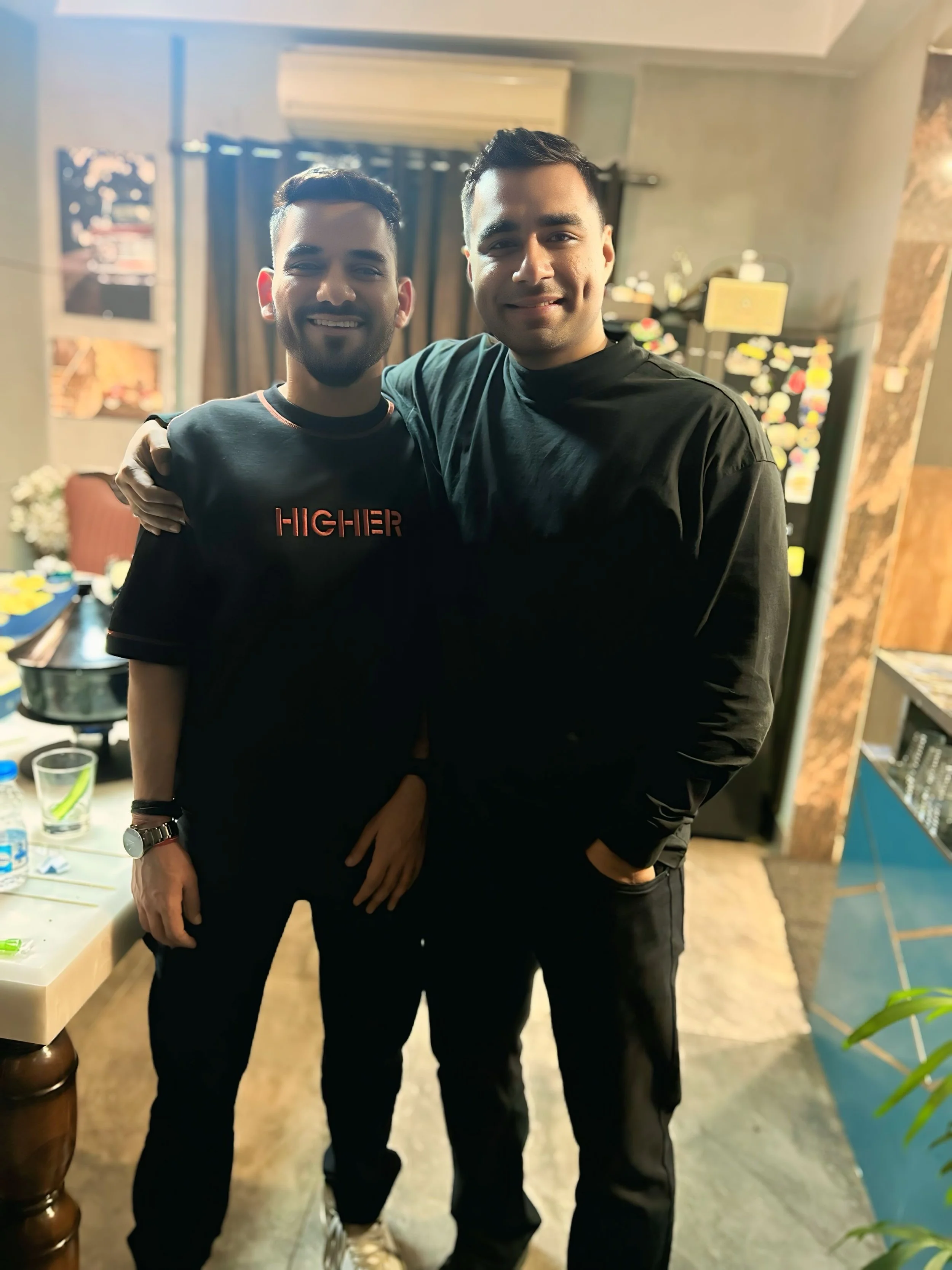 Two men smiling and standing close together indoors, with their arms around each other, in a room with a table, drinks, and a display board in the background.