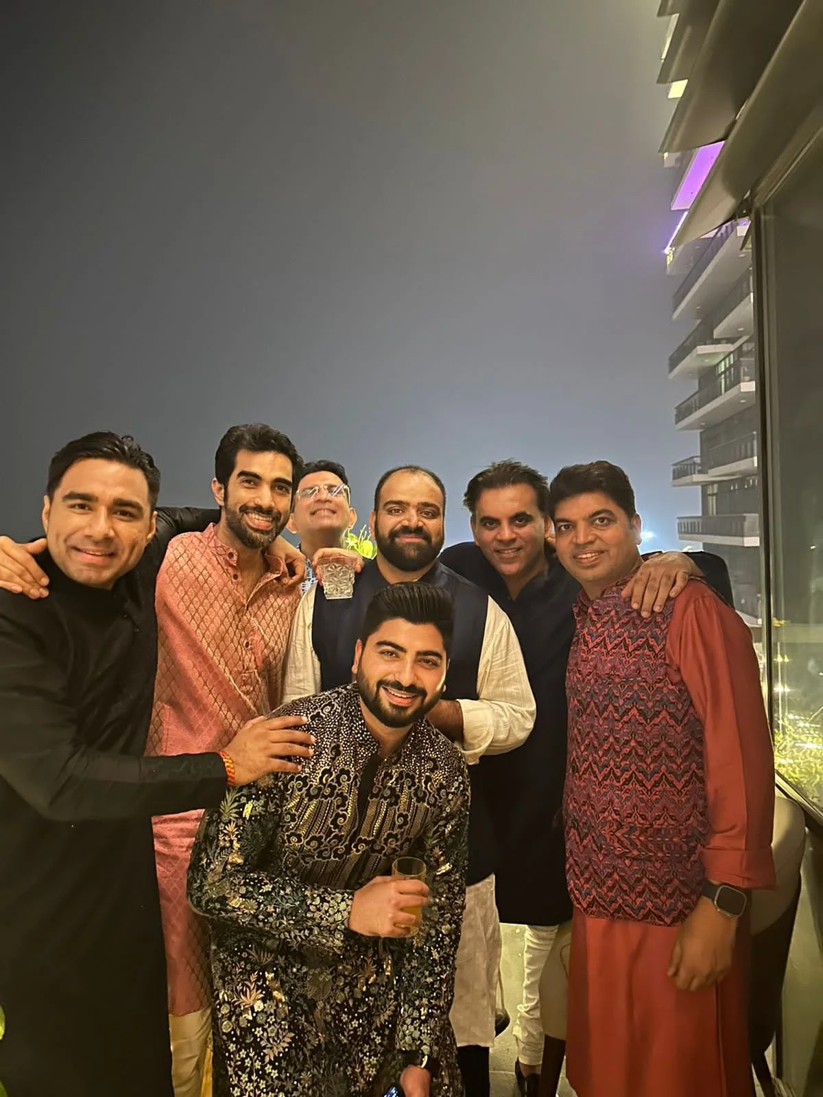 A group of seven men posing together at a night event, some holding drinks, on a balcony with city lights in the background.