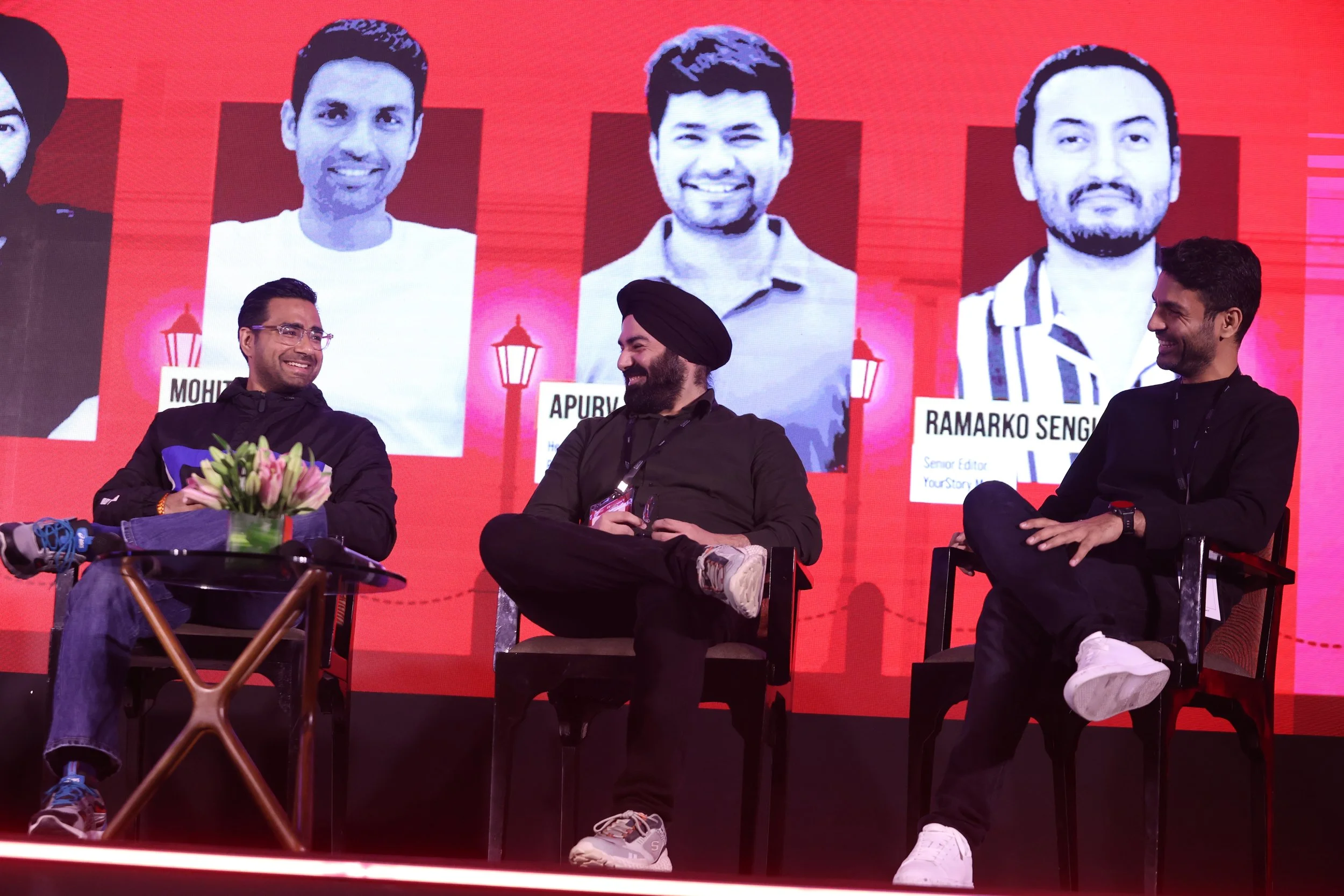 Three men sitting on stage, smiling and engaging in conversation, with a large digital screen behind them displaying photos and names of other individuals.