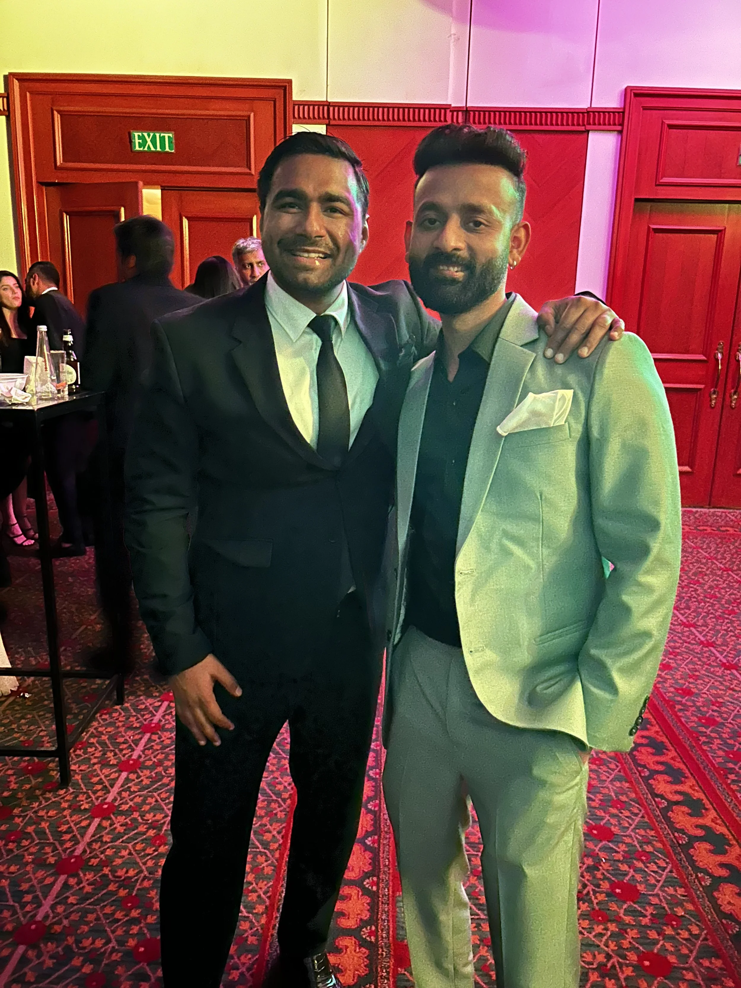 Two men in suits posing together at a formal event, with one wearing a black suit and the other in a light-colored blazer, surrounded by people and red wood paneling.