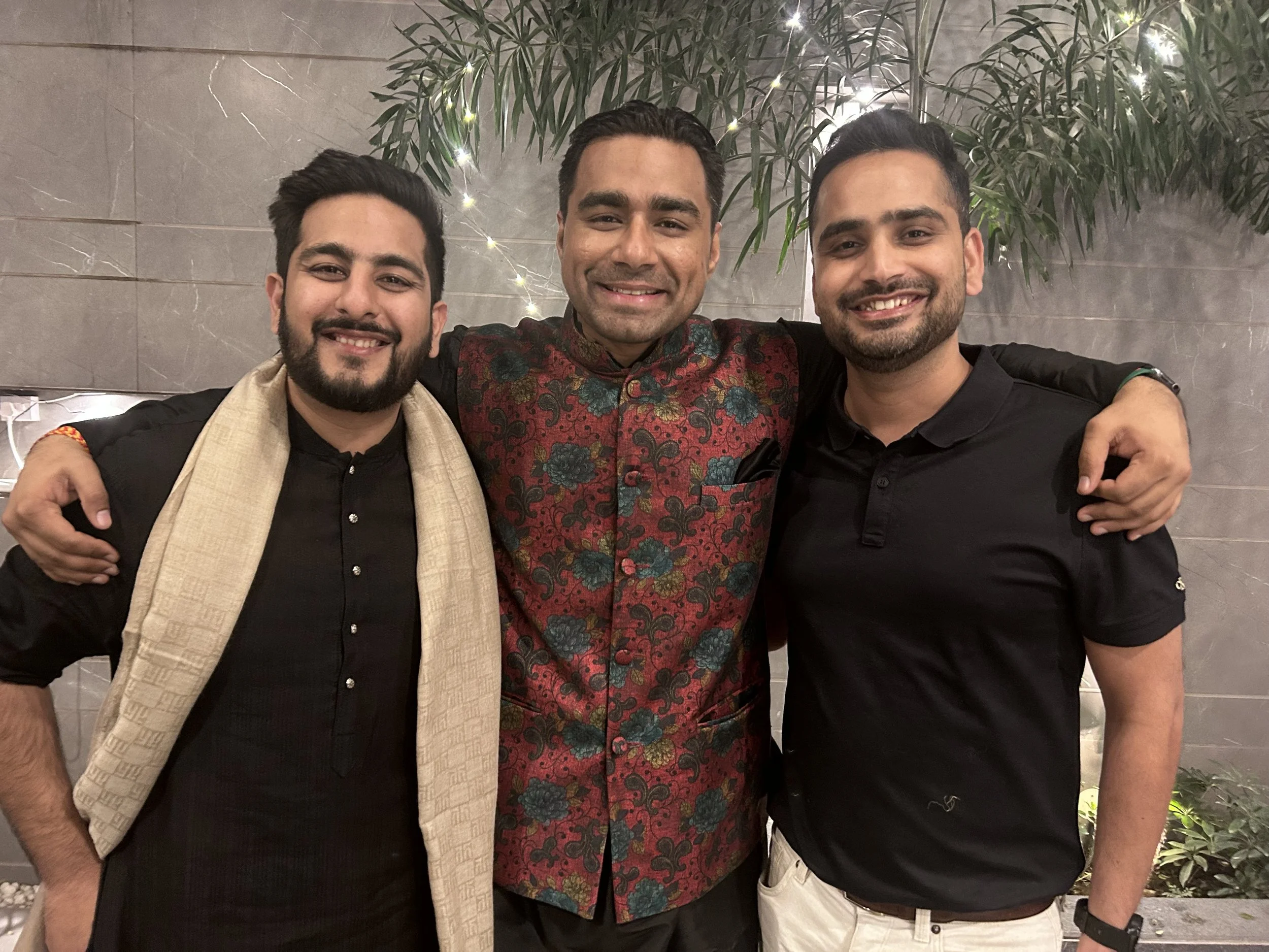 Three men smiling and embracing each other in a festive setting with plants and string lights.
