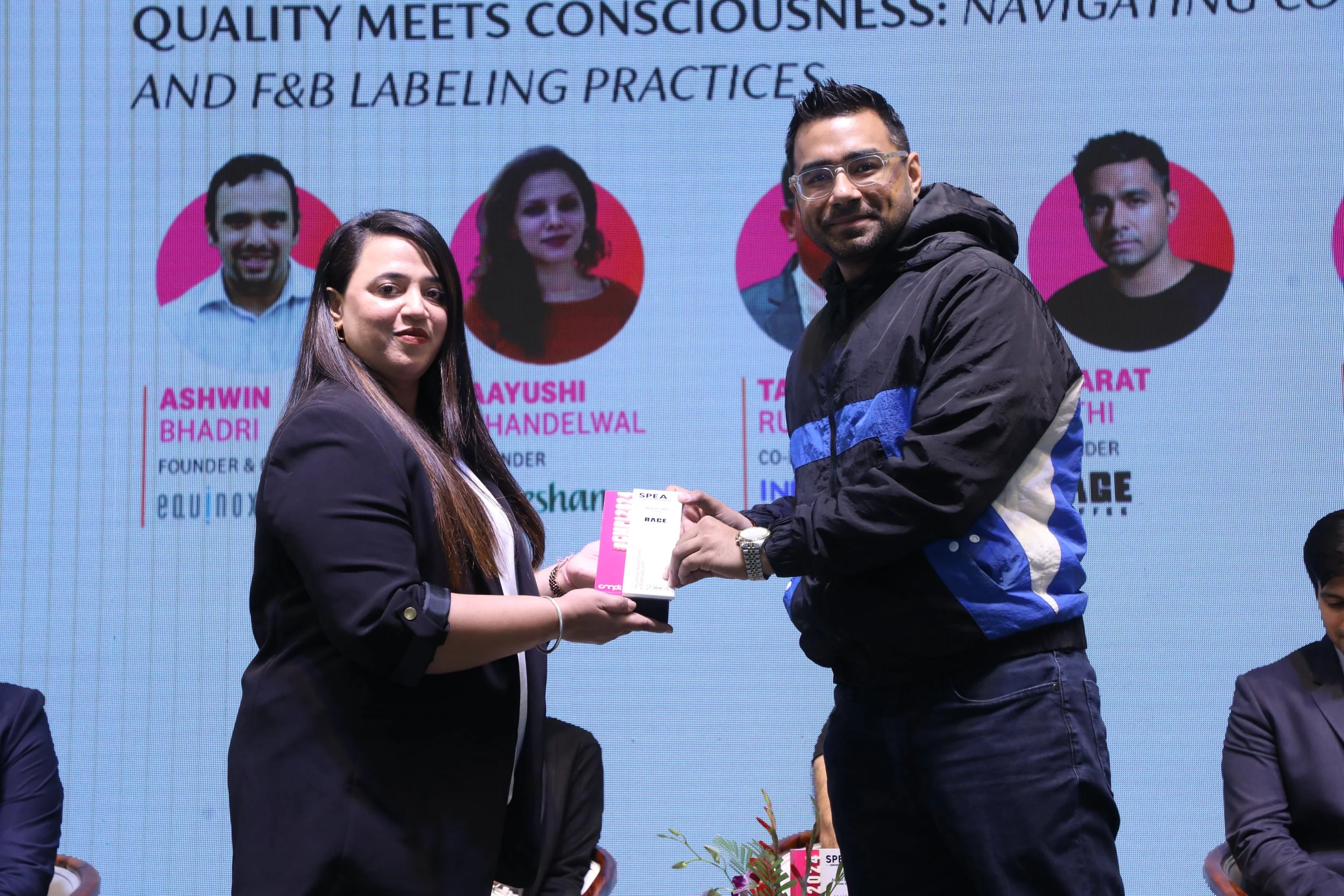 A woman and a man are on stage during an award ceremony. The woman is handing the man a rectangular award. The background features a large screen displaying images and names of speakers or panelists, including Ashwin Bhadri, Mayushi Handelwal, Taran 