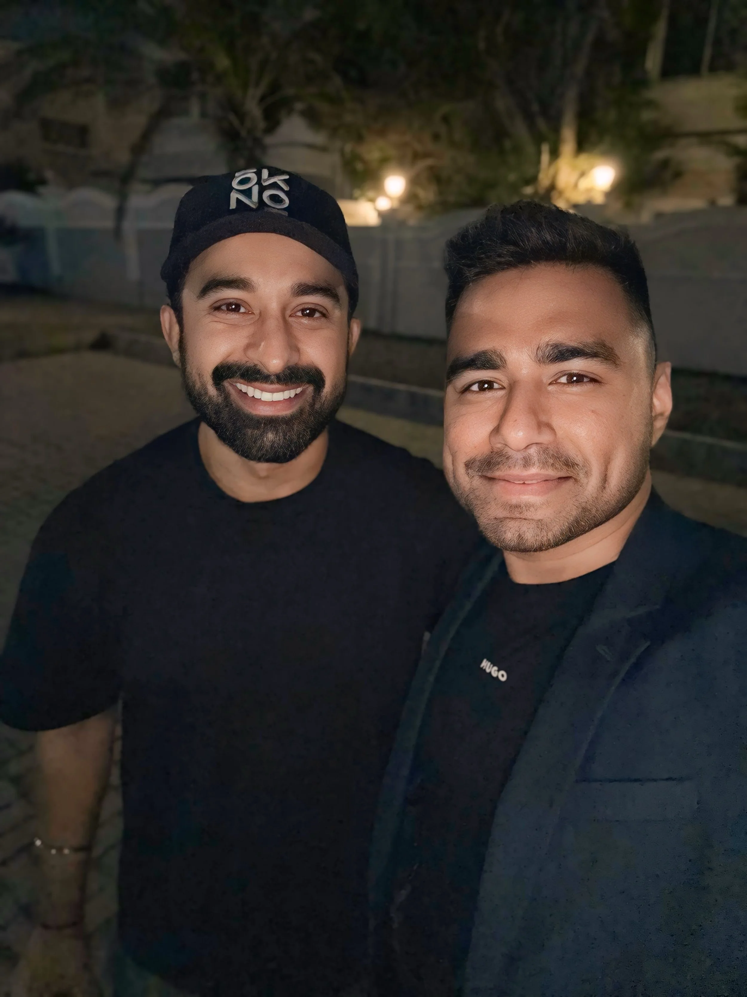 Two men smiling for a selfie outdoors at night, with a dark background and some lights and trees visible.