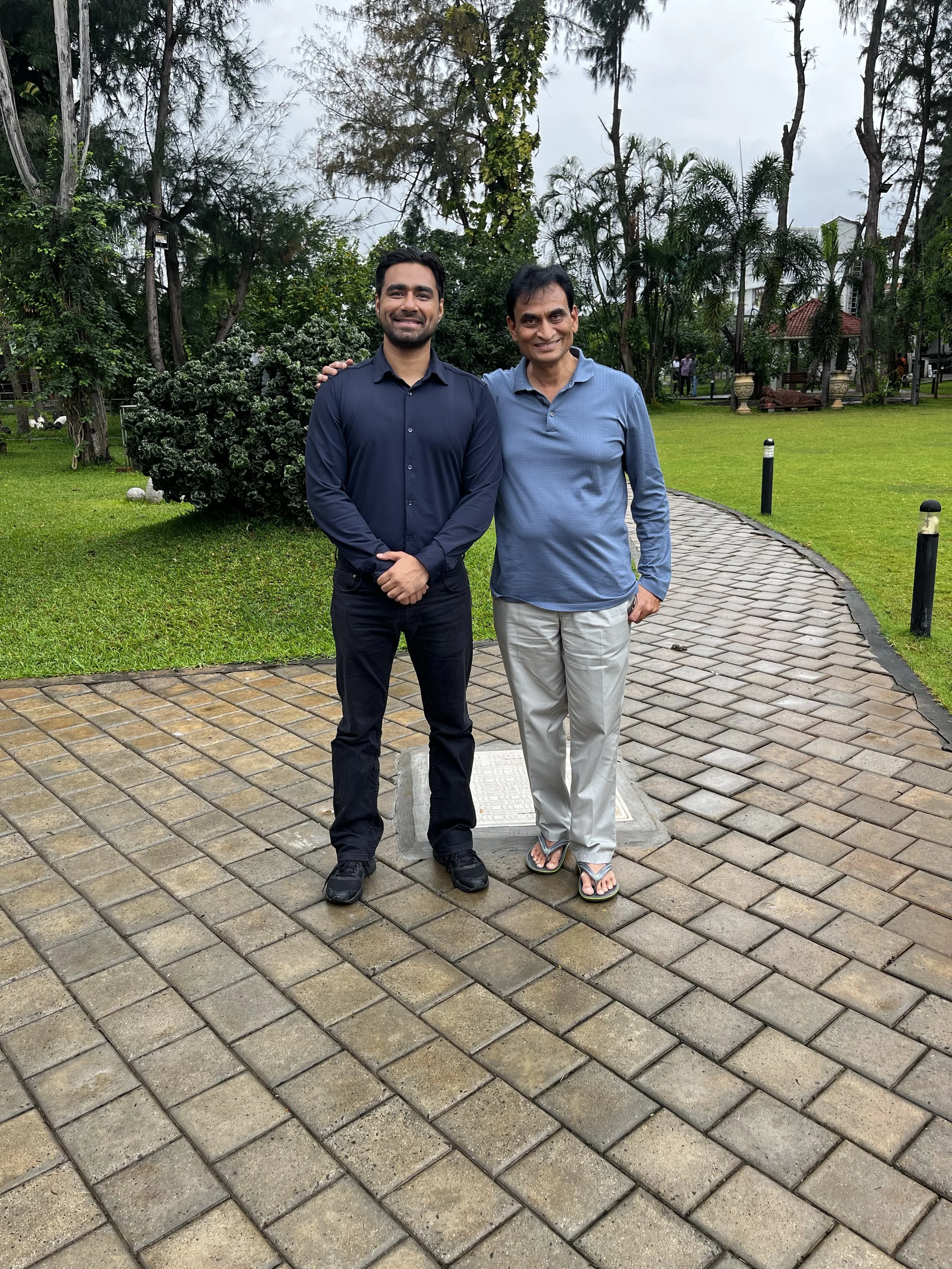 Two men standing on a brick pathway in a park, smiling, with trees and greenery in the background on a cloudy day.