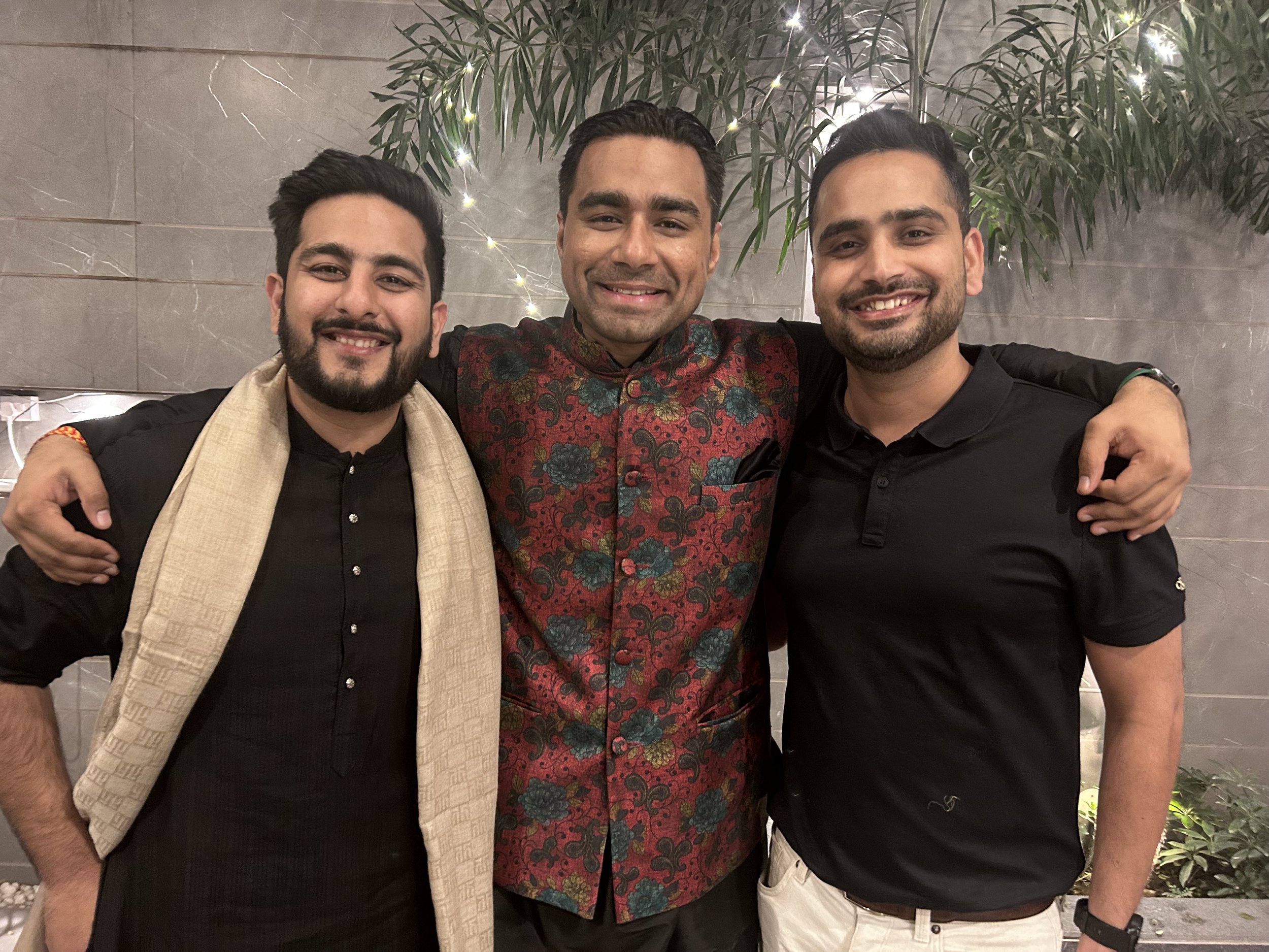 Three men with dark hair and beards, smiling and embracing each other, standing against a gray wall and greenery background.