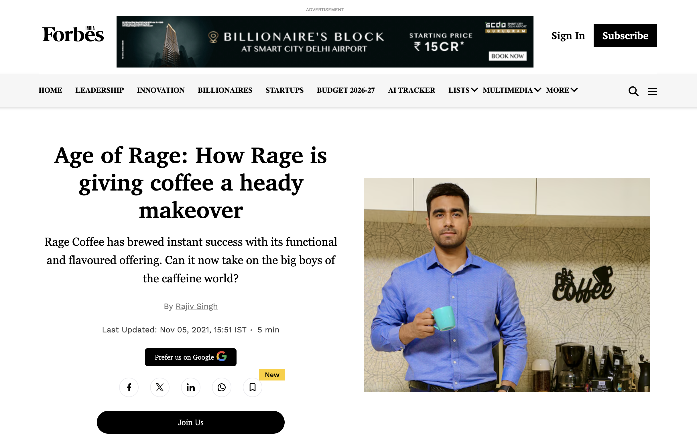 A man in a blue shirt holding a blue coffee mug standing in a coffee shop with a wall behind him that has a coffee-related sign. There are coffee machines on the counter to his right.