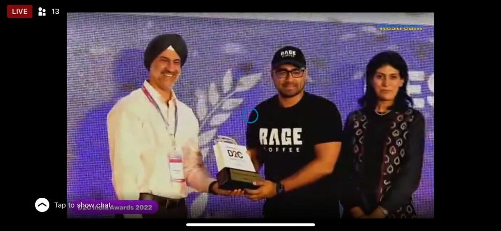Three people standing on a stage, one man in a white shirt and turban holding an award, another man in a black RAGE Coffee T-shirt and cap, and a woman in a black patterned dress. They are participating in an award ceremony at the Spectra Awards 2022