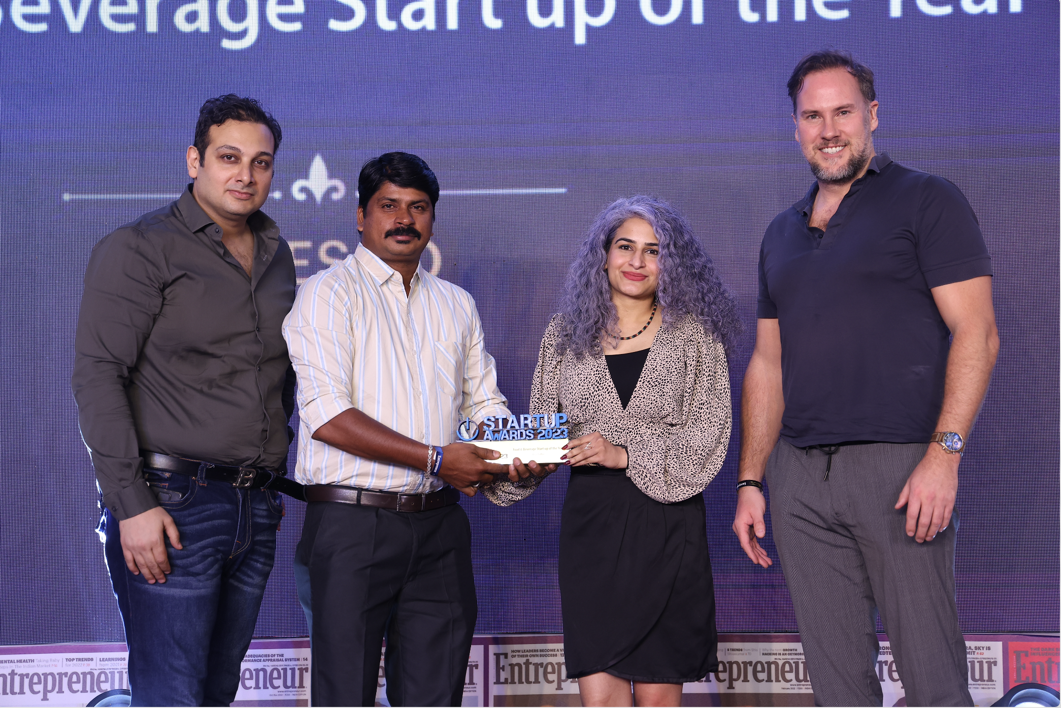 A group of four people standing on stage during an award ceremony, with a backdrop displaying text about startups and awards. One person is holding a trophy that says 'STARTUP AWARDS 2023'.