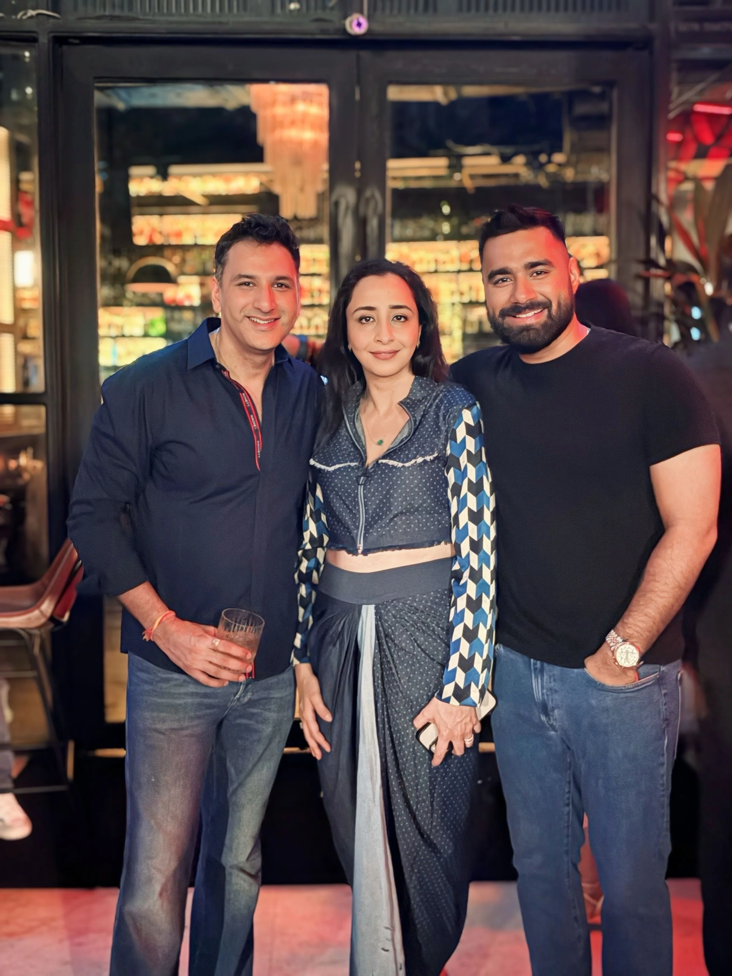 Three people standing together in a bar or restaurant, smiling at the camera. The man on the left is holding a drink, wearing a navy blue shirt and jeans. The woman in the middle has shoulder-length hair, wearing a patterned jacket and a skirt. The m