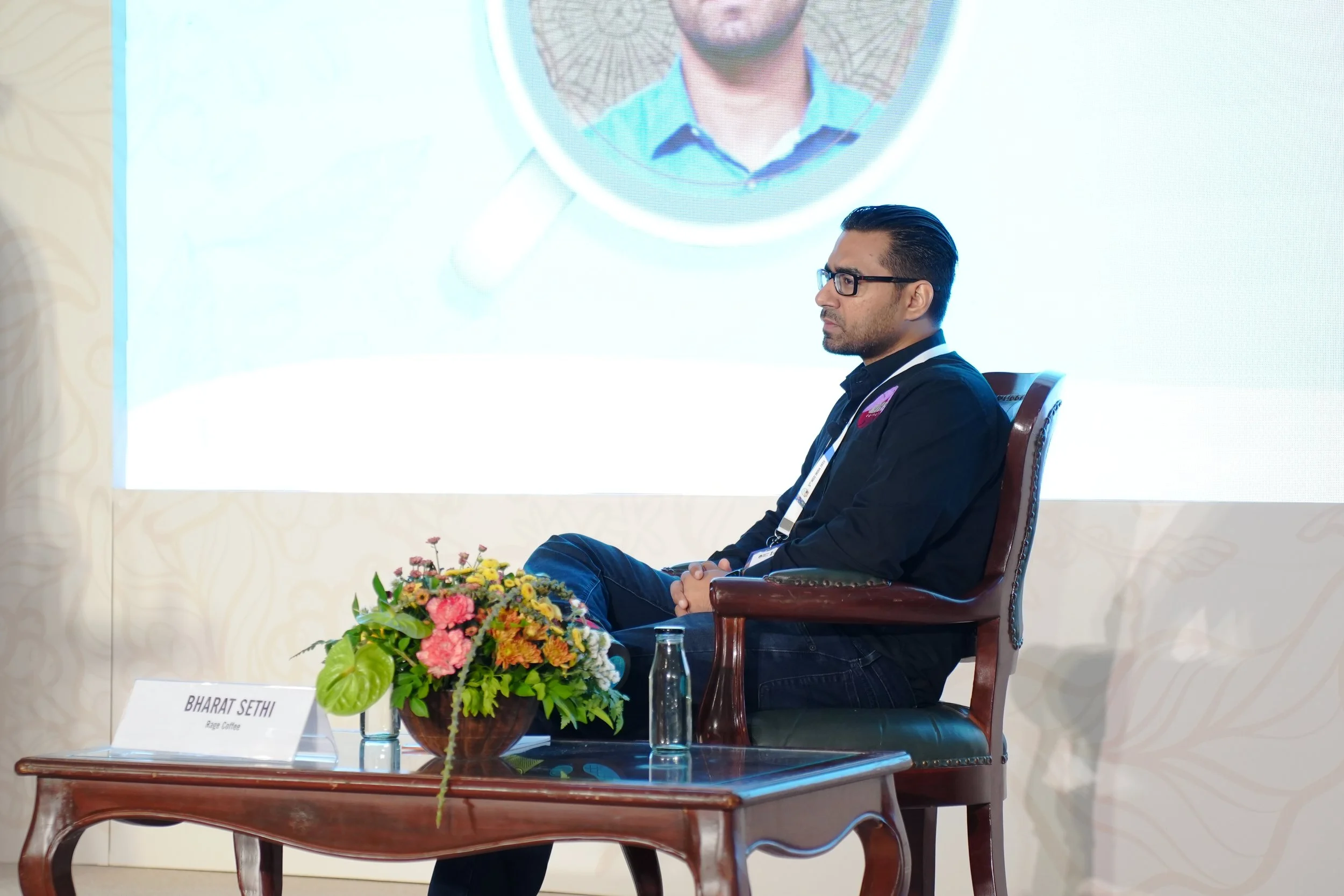 A man with glasses sits on a wooden chair on a stage with a large screen behind him. There is a wooden table in front of him with a bouquet of flowers, a nameplate, and a bottle of water.
