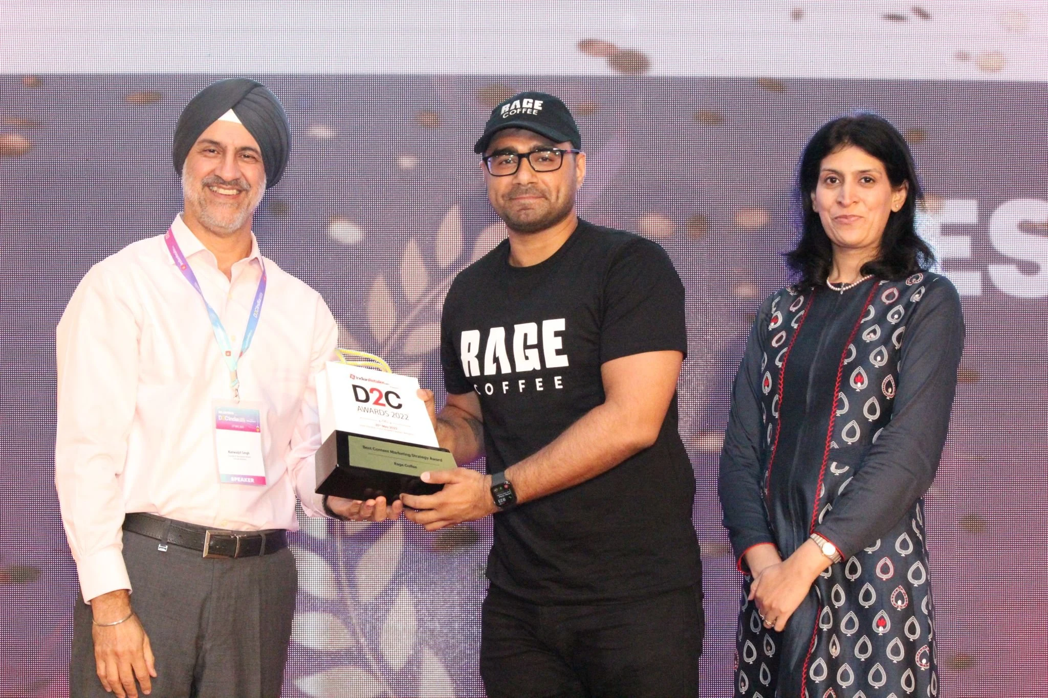 Three people standing on stage at an award ceremony; the man in the middle is holding a trophy. The man on the left is wearing a white shirt and a turban, the man in the center is wearing a black T-shirt and glasses, and the woman on the right is wea