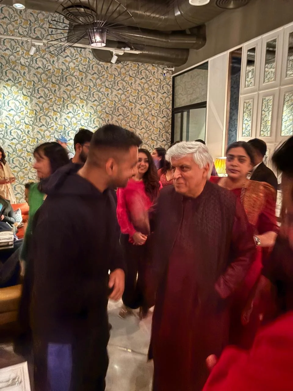 A group of people, mostly women in colorful traditional Indian clothing and men, gather in a warmly lit room with patterned wallpaper, chatting and smiling. An older man with white hair wears a dark traditional outfit and is engaged in conversation w