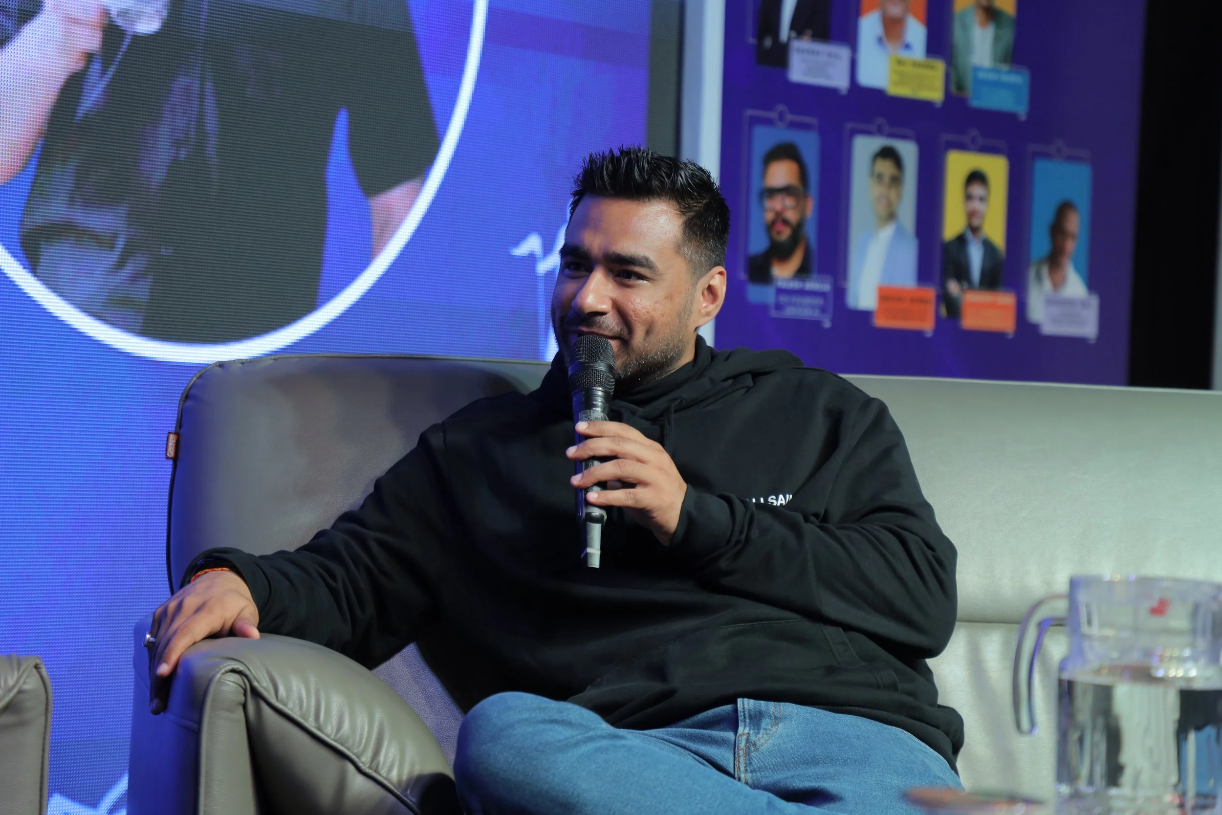 A man with short dark hair and beard, sitting on a gray armchair, holding a microphone, speaking. He is wearing a black hoodie and blue jeans. Behind him, a screen displays multiple photos and info of people, with a purple background.