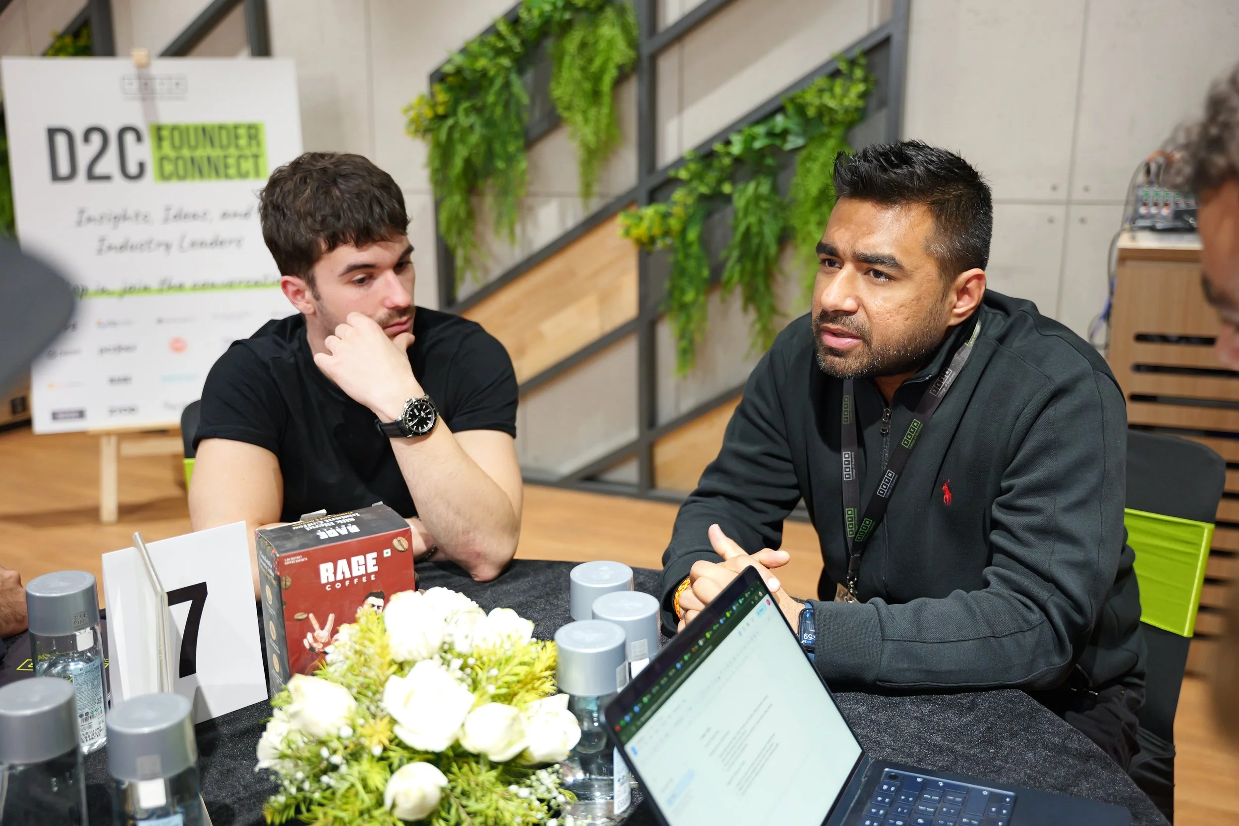 Two men sitting at a table engaged in conversation, with a laptop, bottles of hand sanitizer, a coffee box, and a flower arrangement on the table, in a modern indoor space with green plants on the wall.