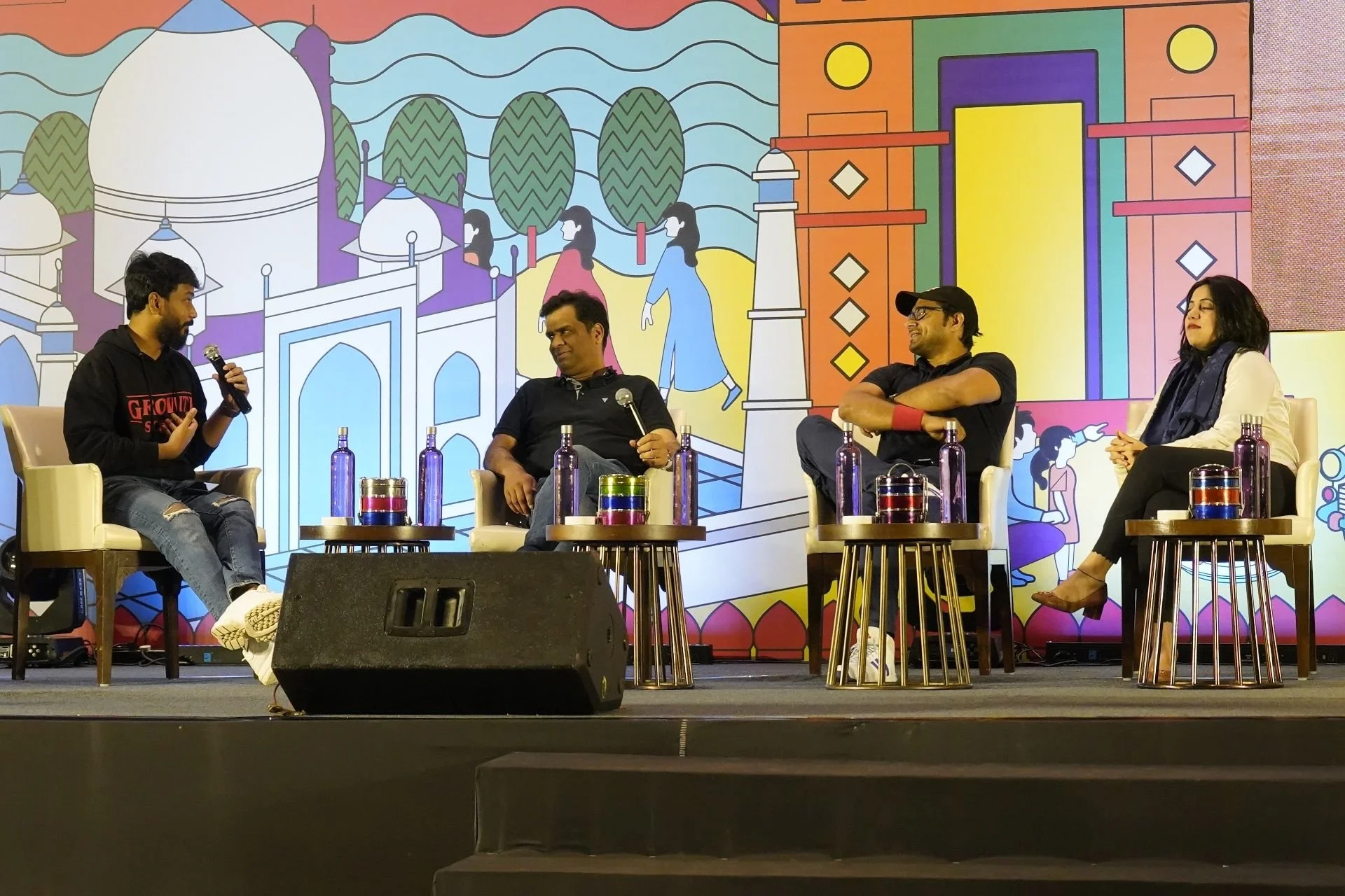 Four people sitting on stage having a discussion, with colorful backdrops featuring traditional Indian architecture and figures.