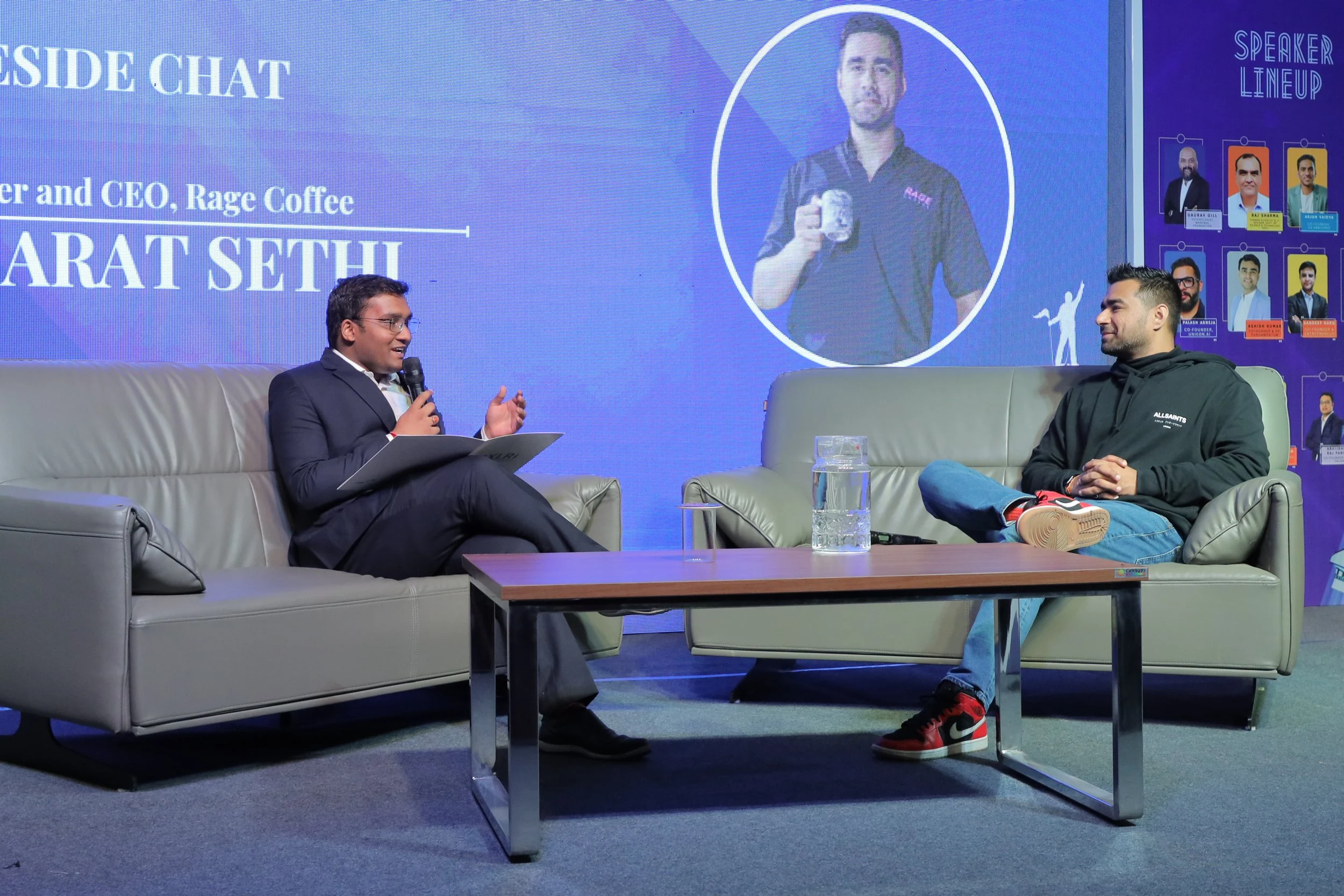 Two men having a discussion on stage, seated on modern gray sofas, with a wooden coffee table in between. The man on the left, dressed in a suit, is speaking into a microphone and holding a folder. The man on the right, in casual attire including a b