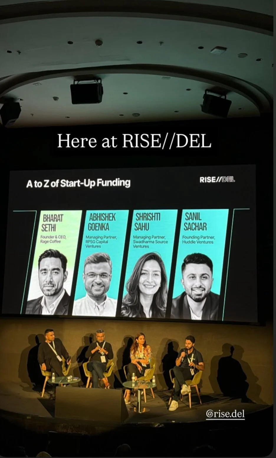 Panel discussion at RISE//DEL event, with a large screen displaying profiles of four startup founders and their companies, seated on stage with audience shadows visible.