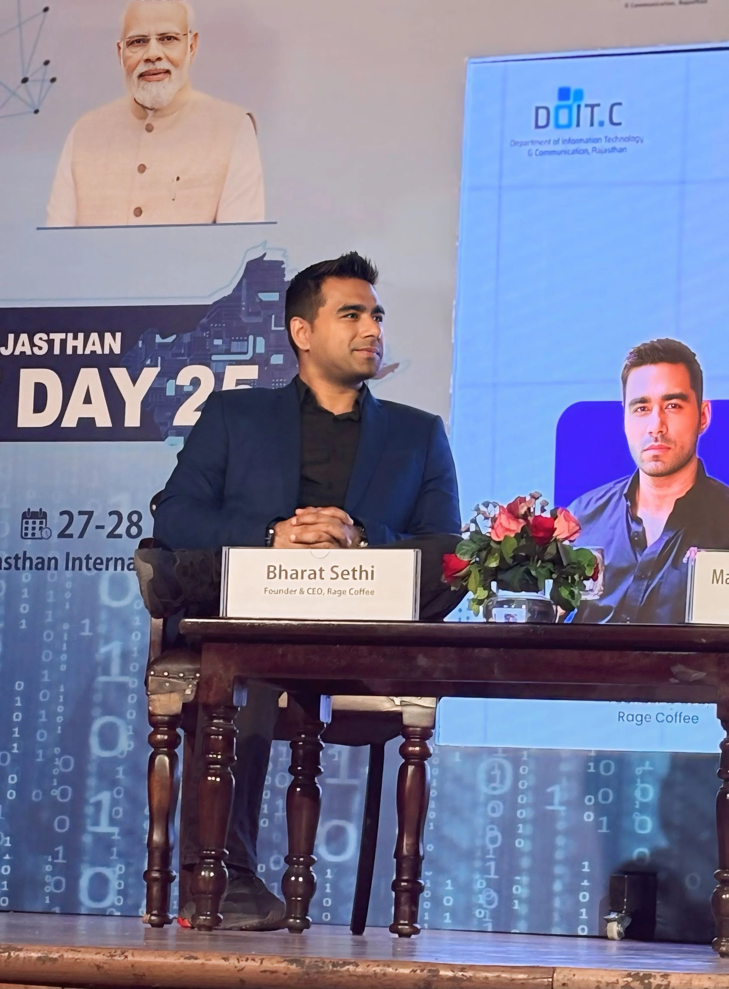 A man in a dark suit sits at a table with a nameplate that reads Bharat Sethi, Founder & CEO of Rage Coffee. Behind him is a large backdrop with text indicating a technology conference. There is a bouquet of flowers on the table and a background with
