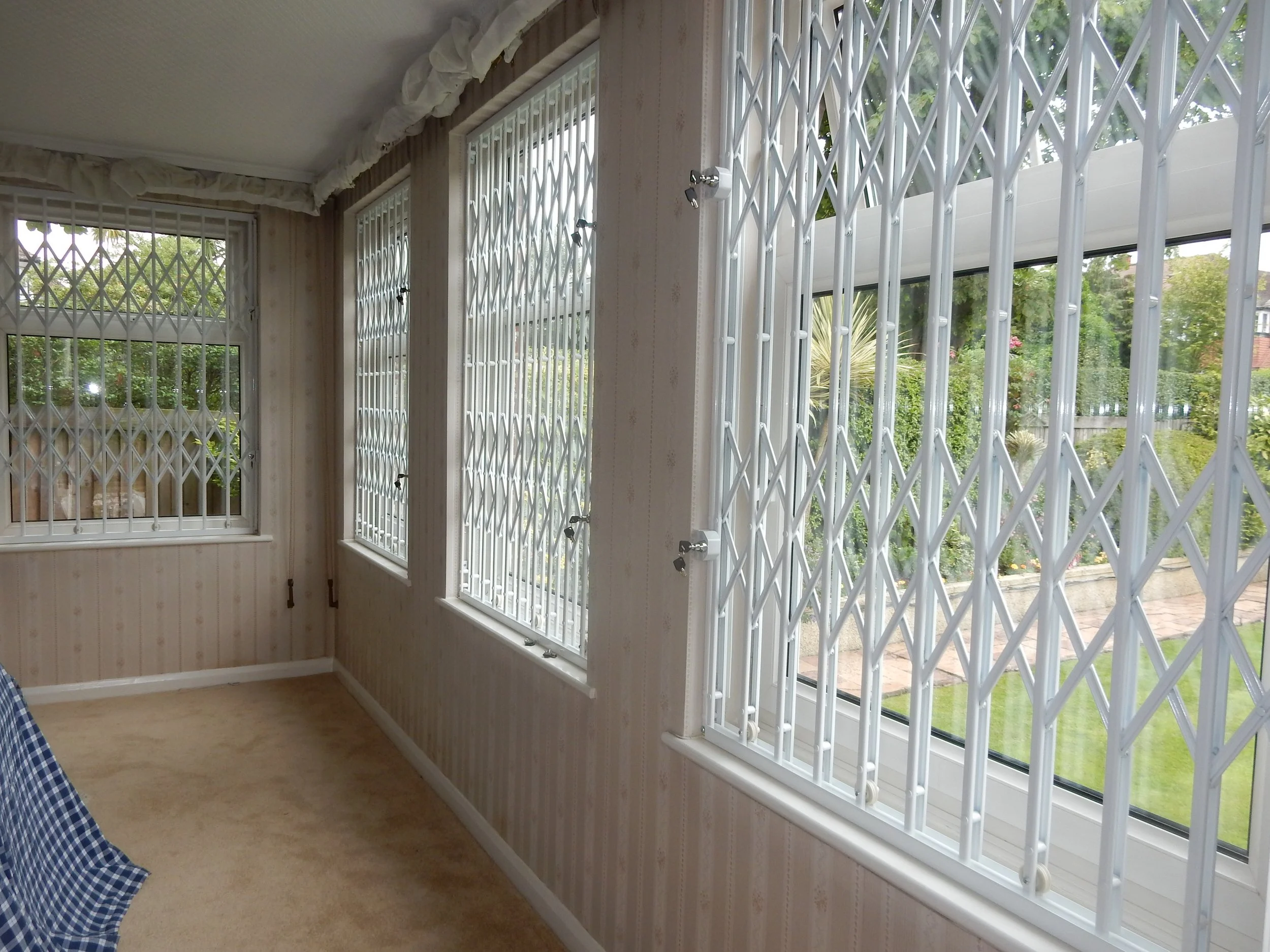 Room with three large windows covered by white security bars, beige patterned wallpaper, and beige carpet. Outside view shows greenery and a brick sidewalk.