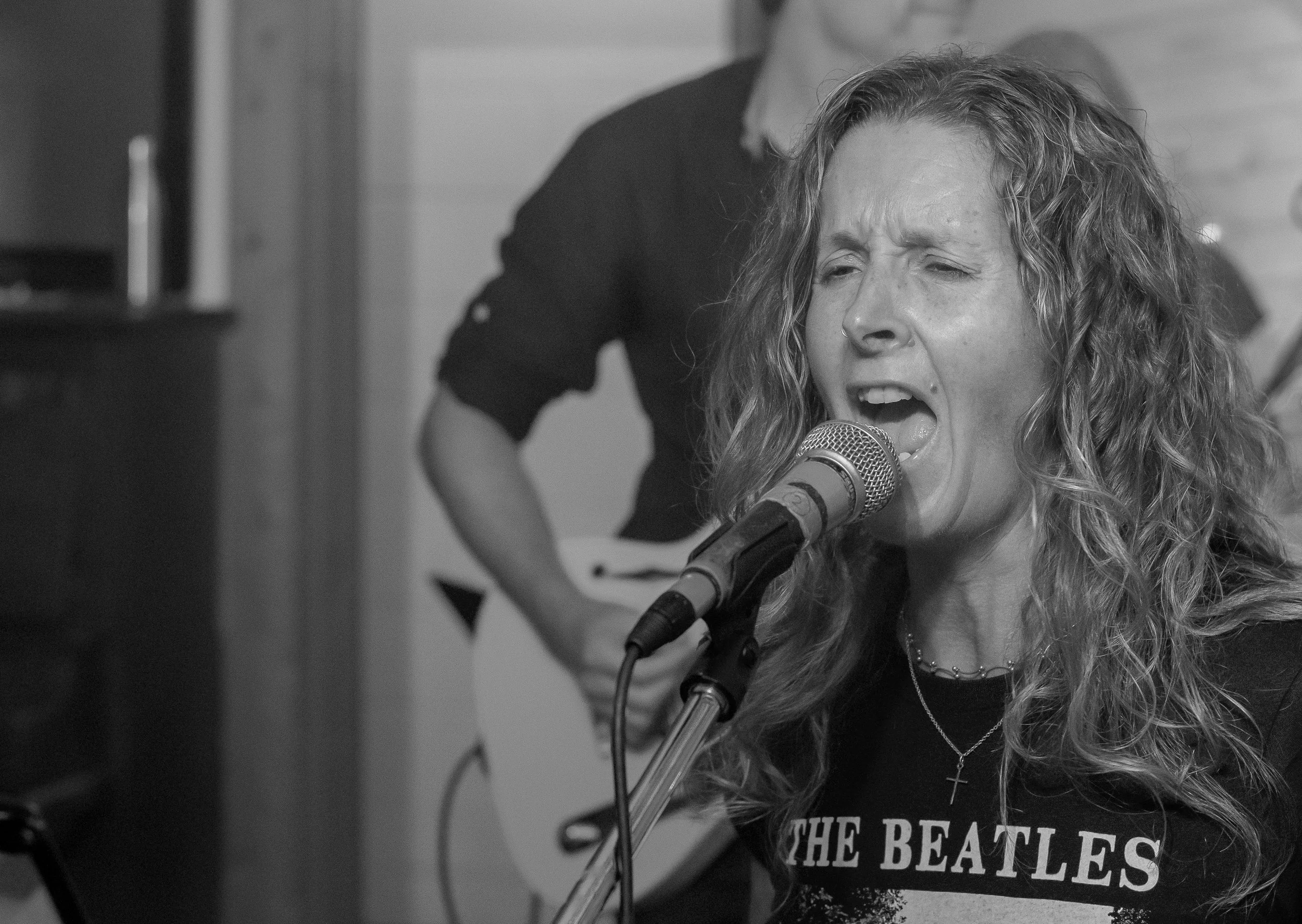 A woman with curly hair singing into a microphone, wearing a 'The Beatles' t-shirt, with a person playing guitar in the background.