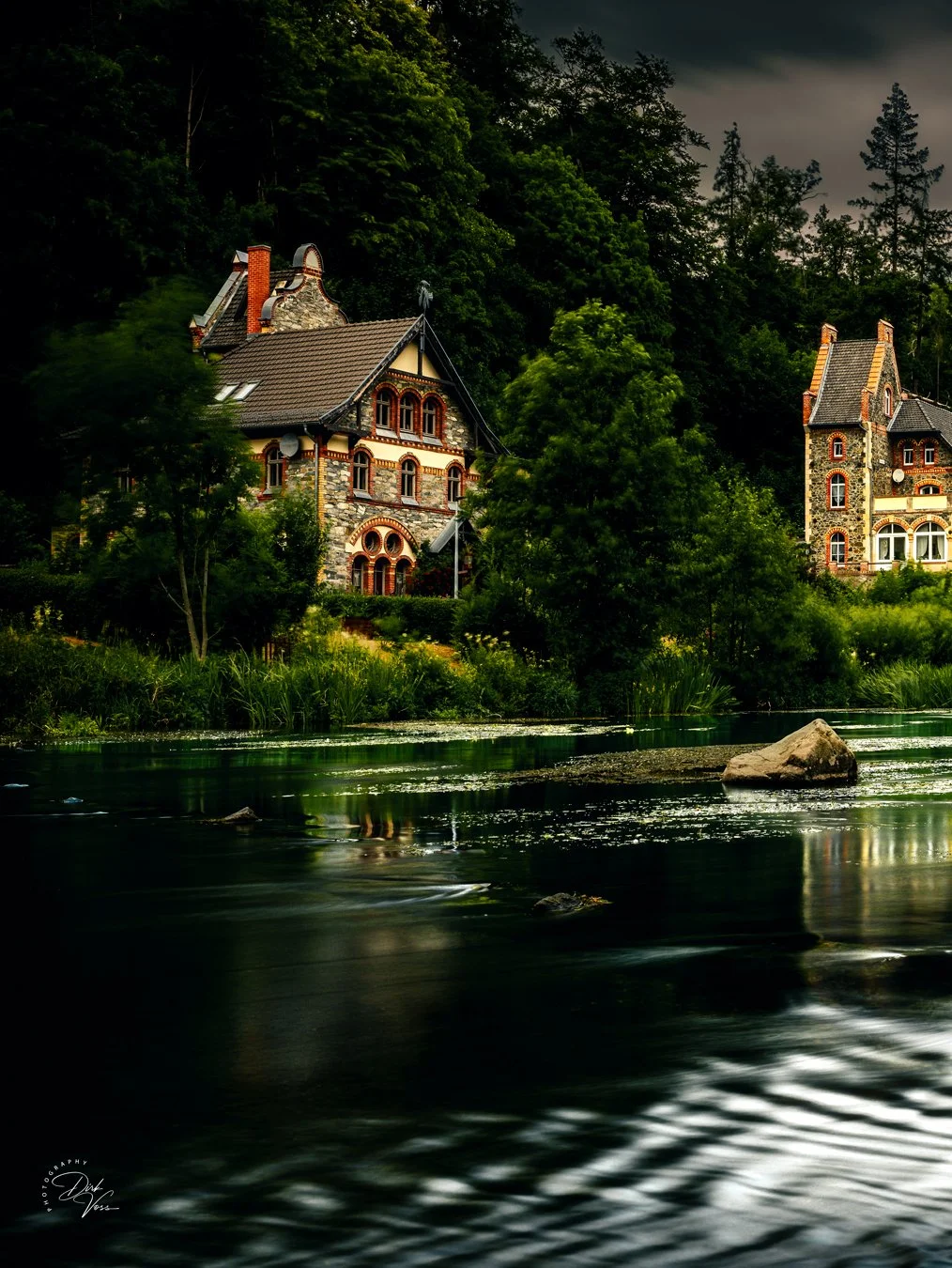 Diese Aufnahme zeigt zwei historische Häuser mit steilen Dächern und bunten Fenstern, die inmitten eines Waldes am Ufer eines Flusses liegen. Das Wasser reflektiert die Bäume und Gebäude, während der Himmel bewölkt ist.