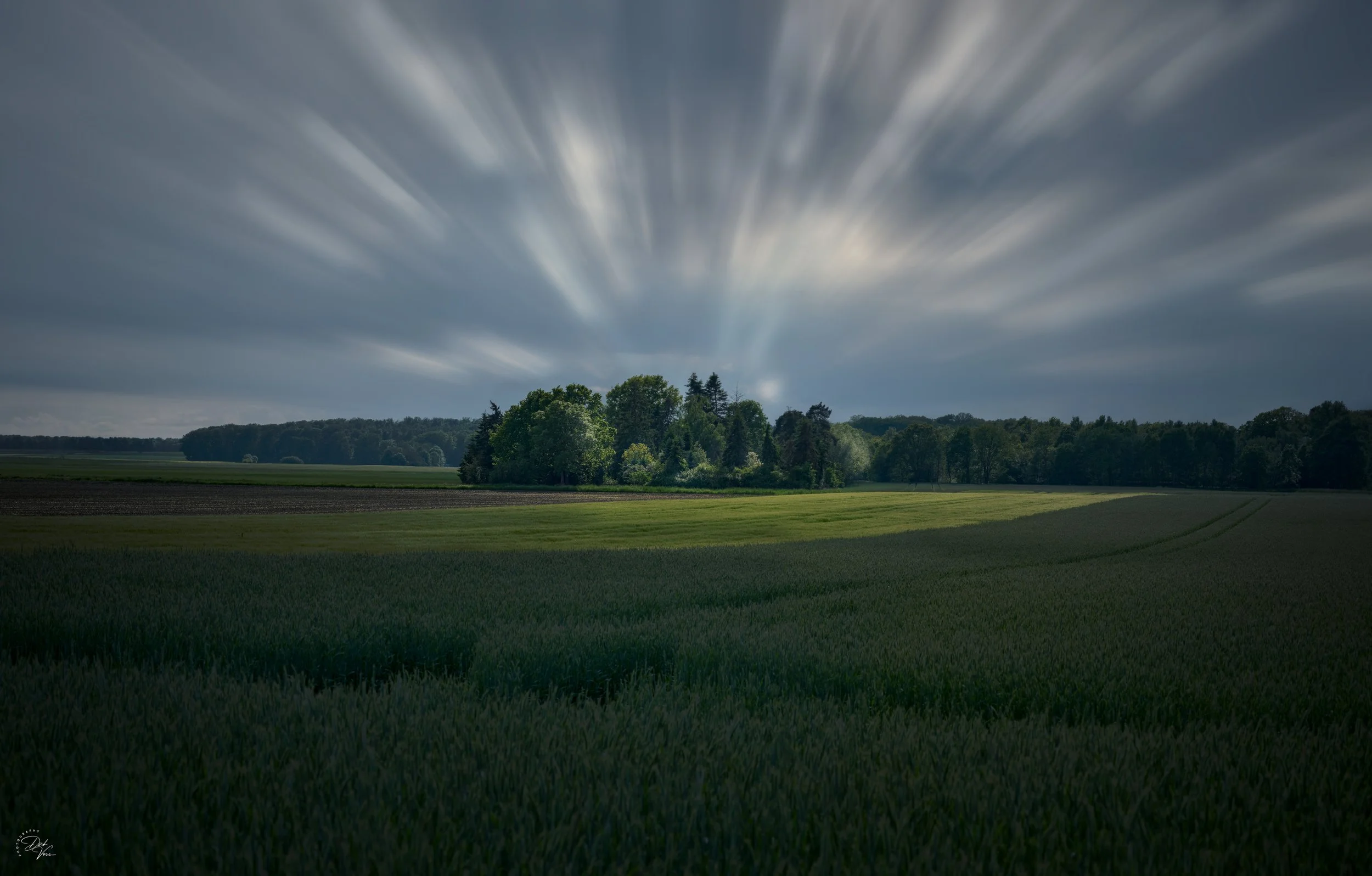 Landschaft mit weiten Feldern, Bäumen und einem bewölkten Himmel mit vorbeiziehenden Wolken.