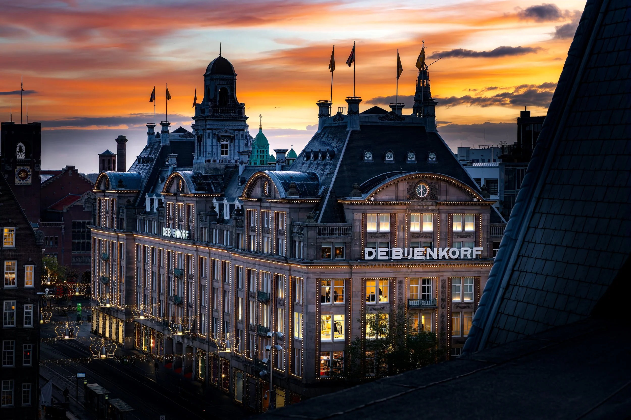 Ein Blick auf die Dächer eines Gebäudes bei Sonnenuntergang mit stilvollen Türmen und Flaggen, die gegen den farbenfrohen Himmel aufragen, darunter das Gebäude mit dem Schriftzug 'DE BUENKORF'.