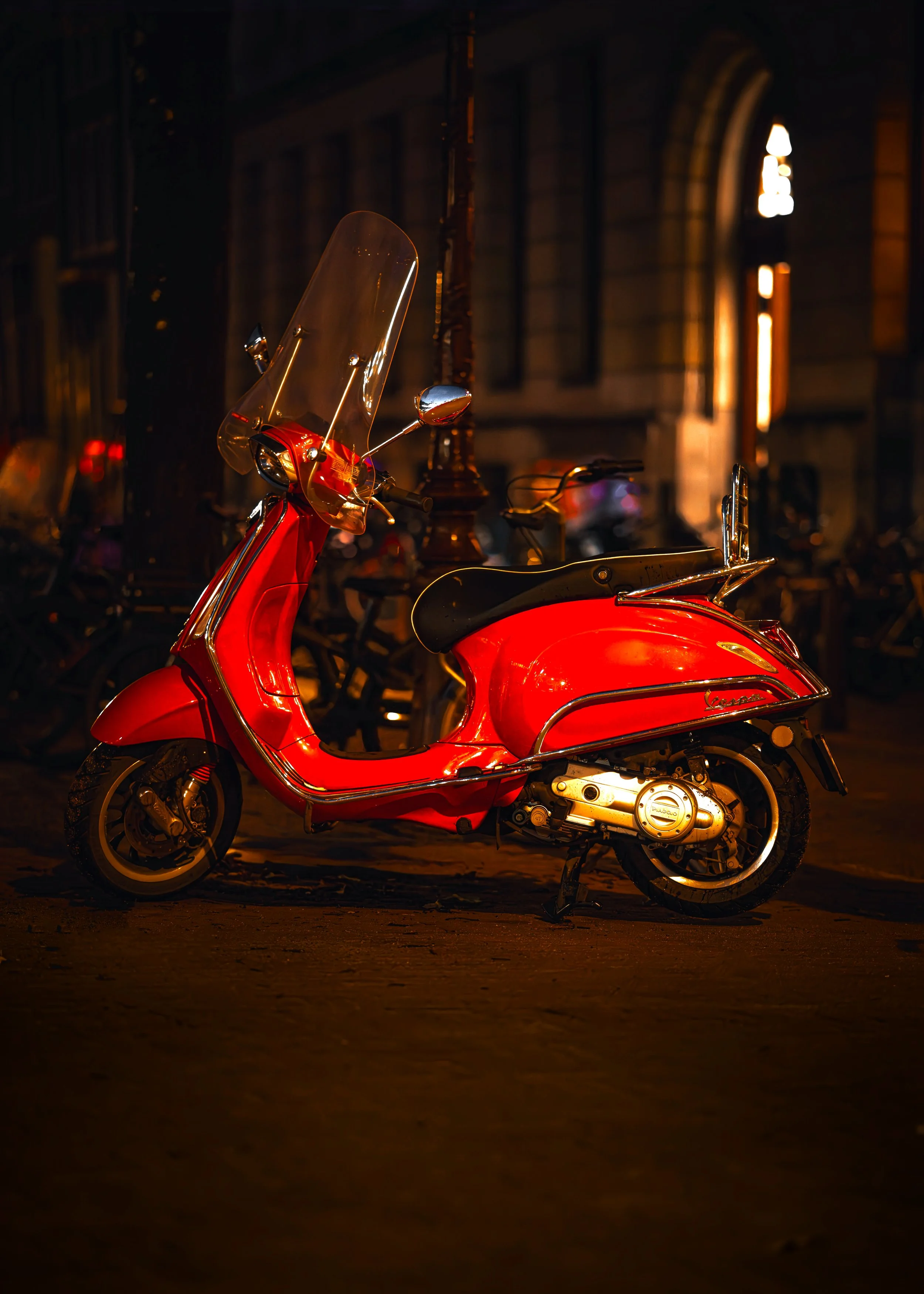 Ein roter Vespa-Roller steht morgens auf einer dunklen Straße, im Hintergrund sind alte Gebäude und Straßenlaternen, die warmes Licht spenden.